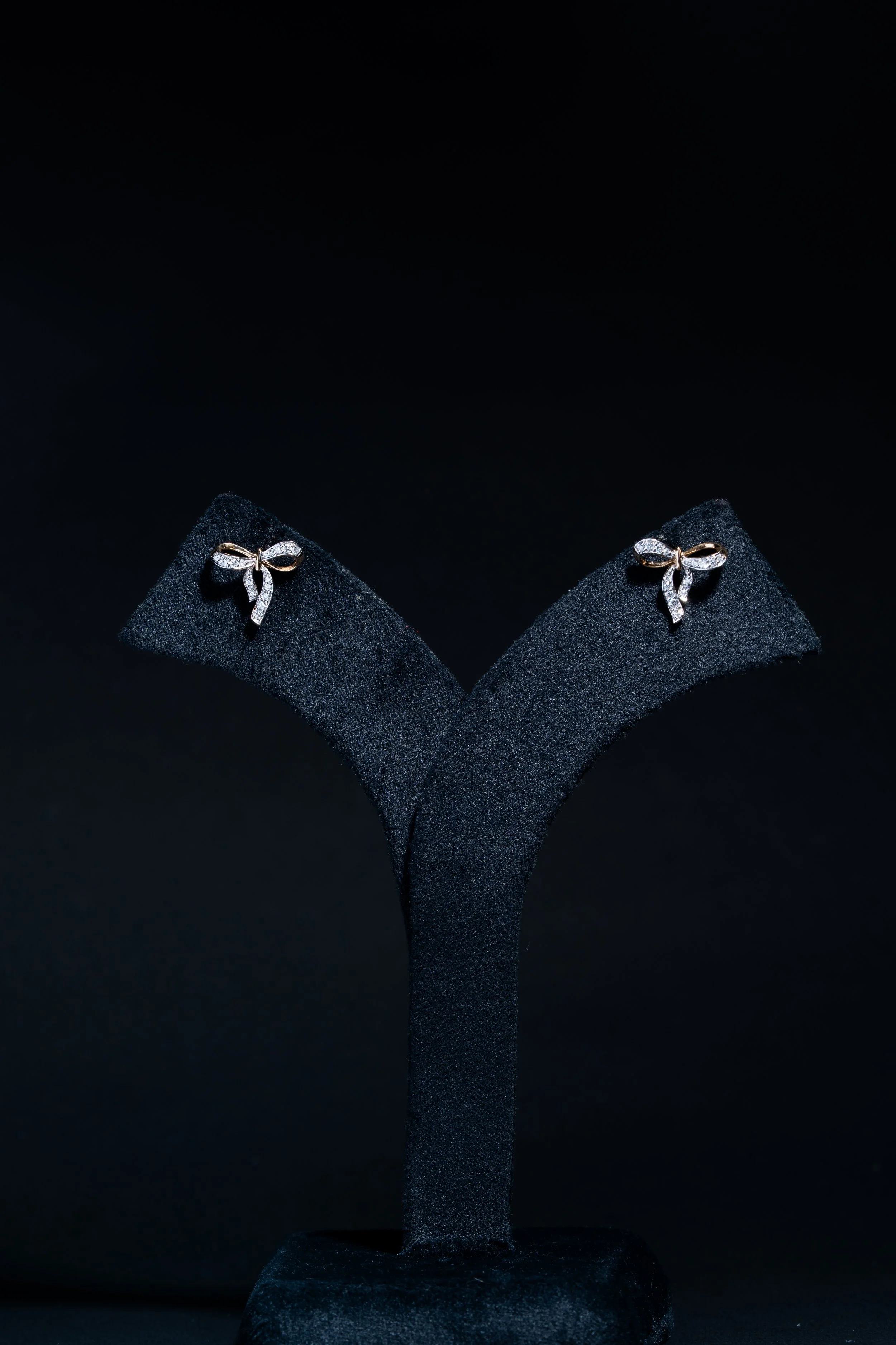 Pair of diamond-studded bow-shaped earrings displayed on black padded stands against a dark background.