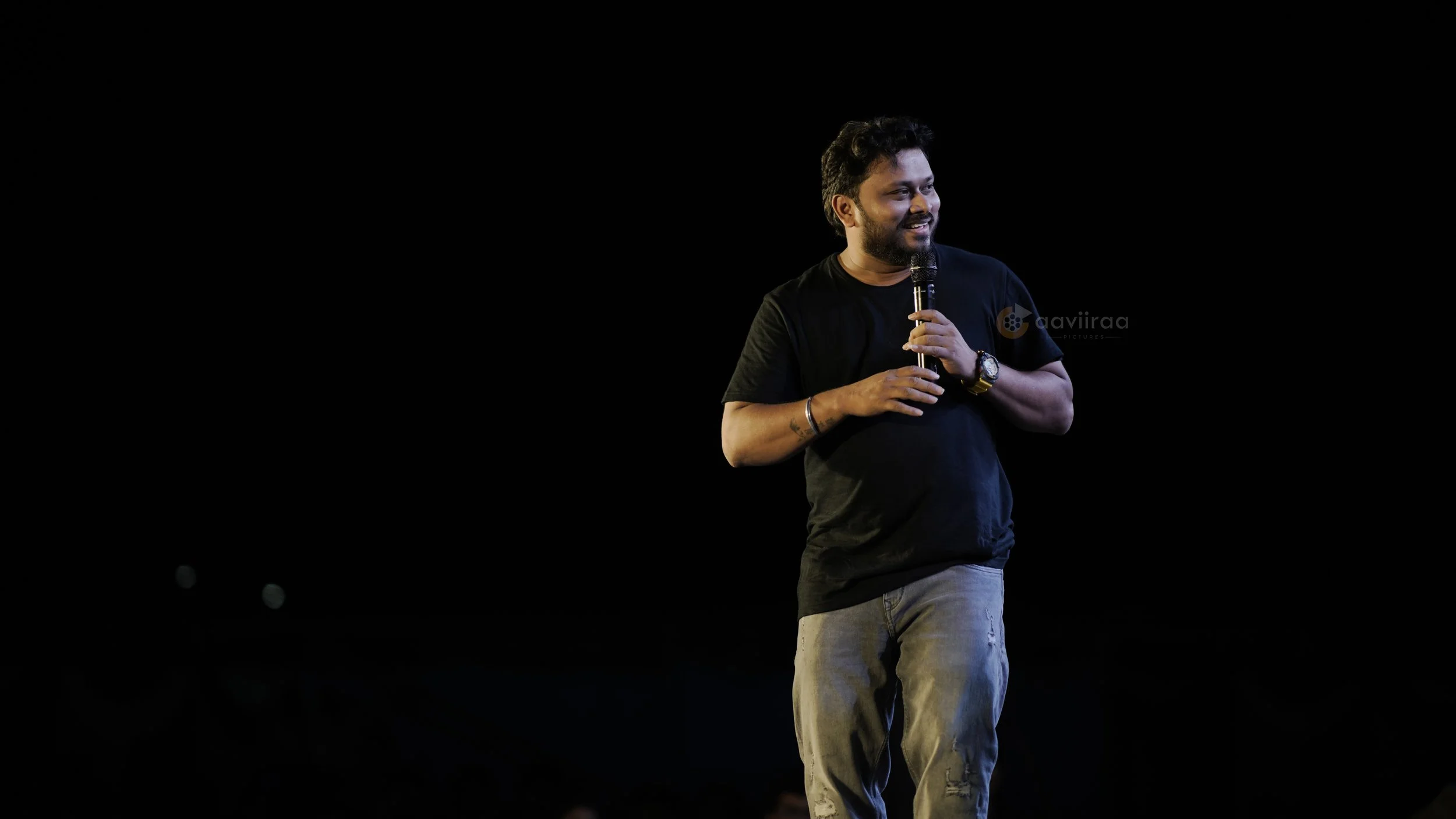A man standing on stage holding a microphone, smiling, wearing a black t-shirt and light-colored pants.
