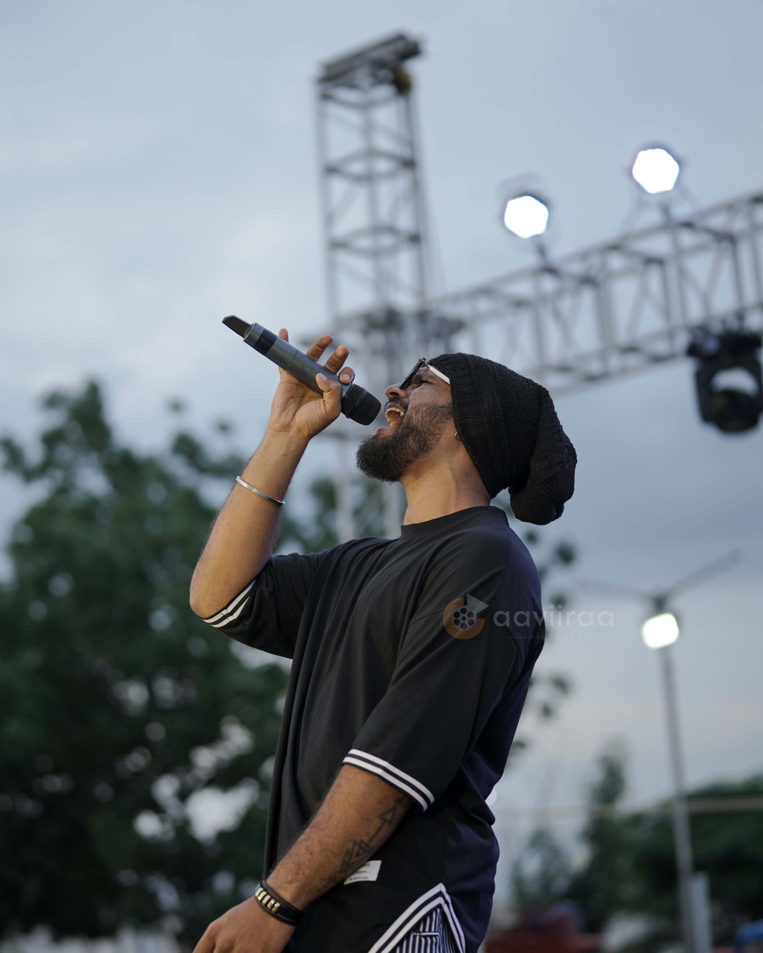 A young man with glasses and a black beanie sings into a microphone on an outdoor stage, with metal truss structures and bright stage lights in the background.