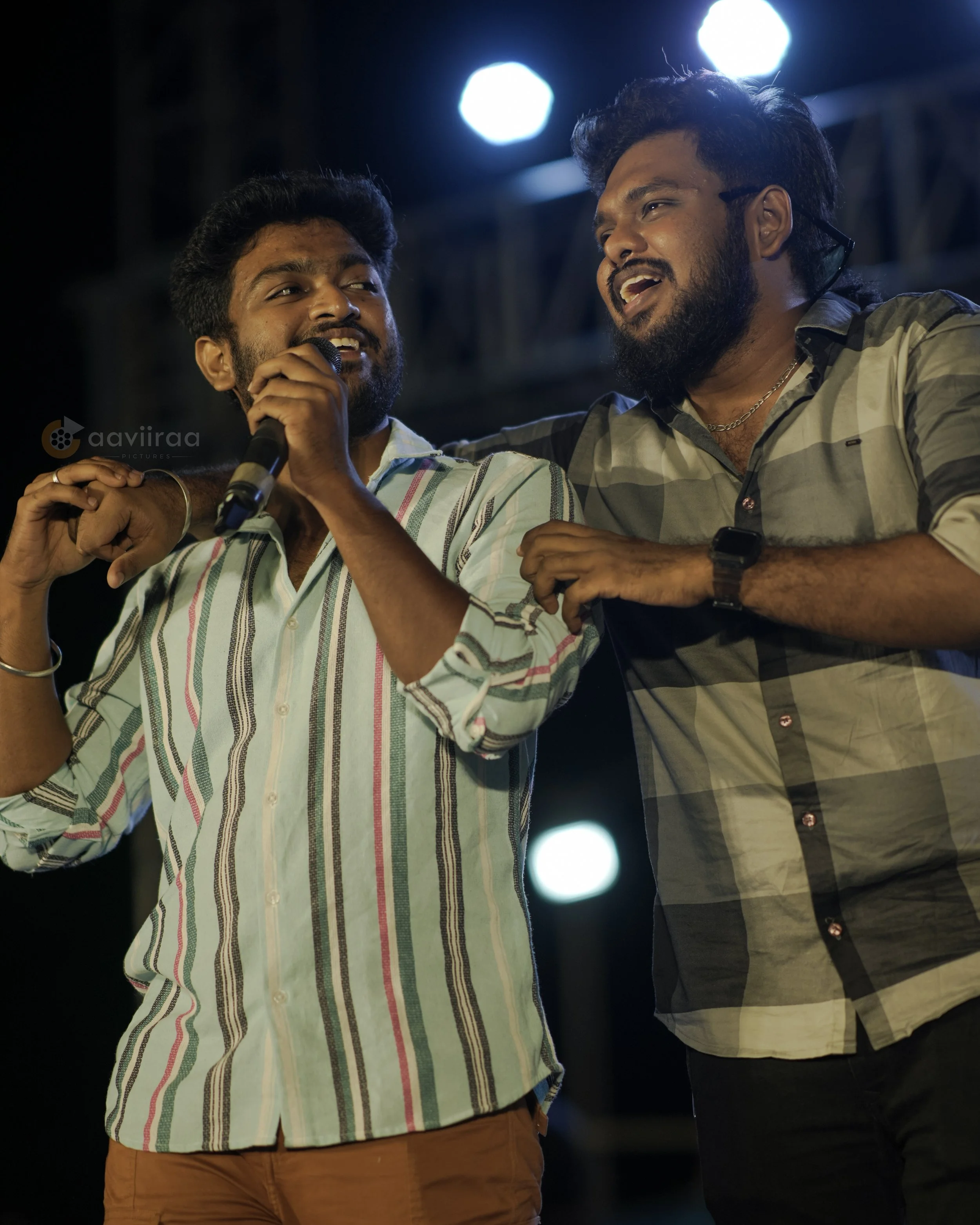 Two men on stage, one holding a microphone, smiling and singing together with bright stage lights overhead.