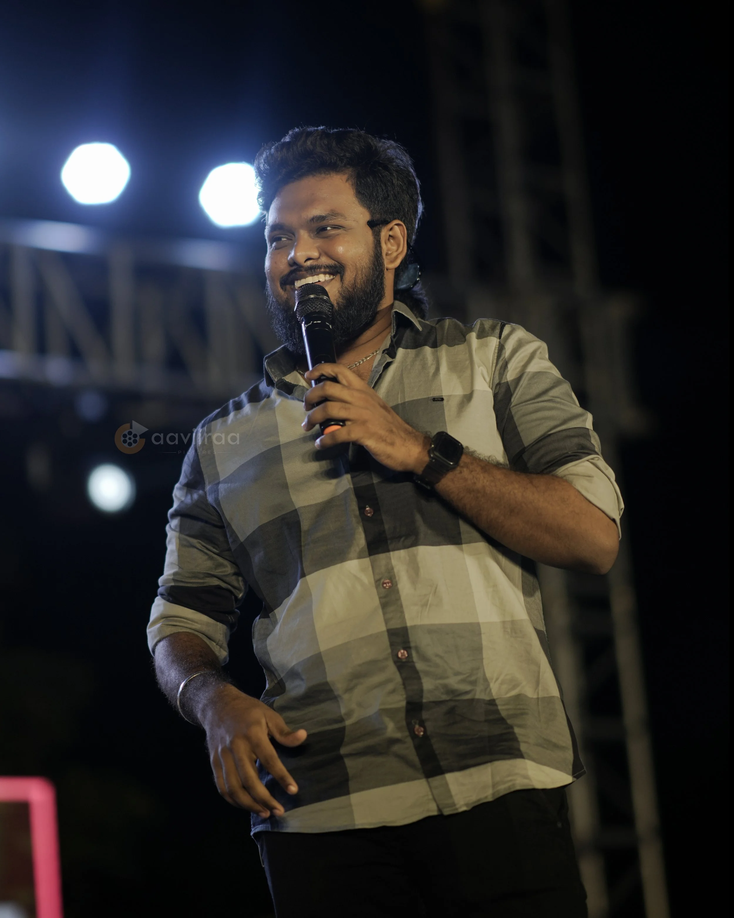 A man with dark hair and beard smiling while holding a microphone, wearing a plaid shirt, standing outdoors at night with bright lights in the background.