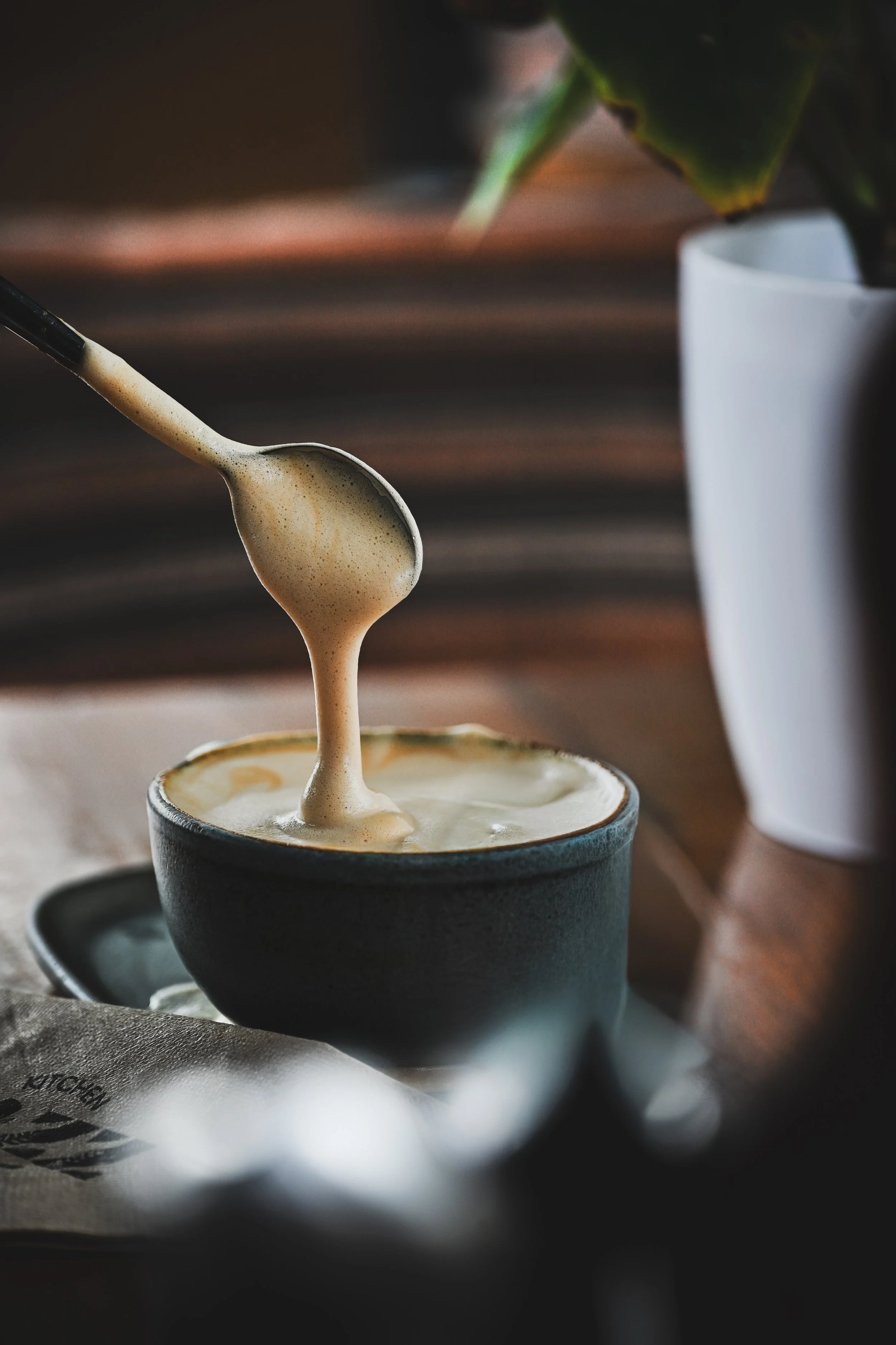 A spoon drizzling frothy coffee into a black ceramic cup, with a white mug and a plant in the background.