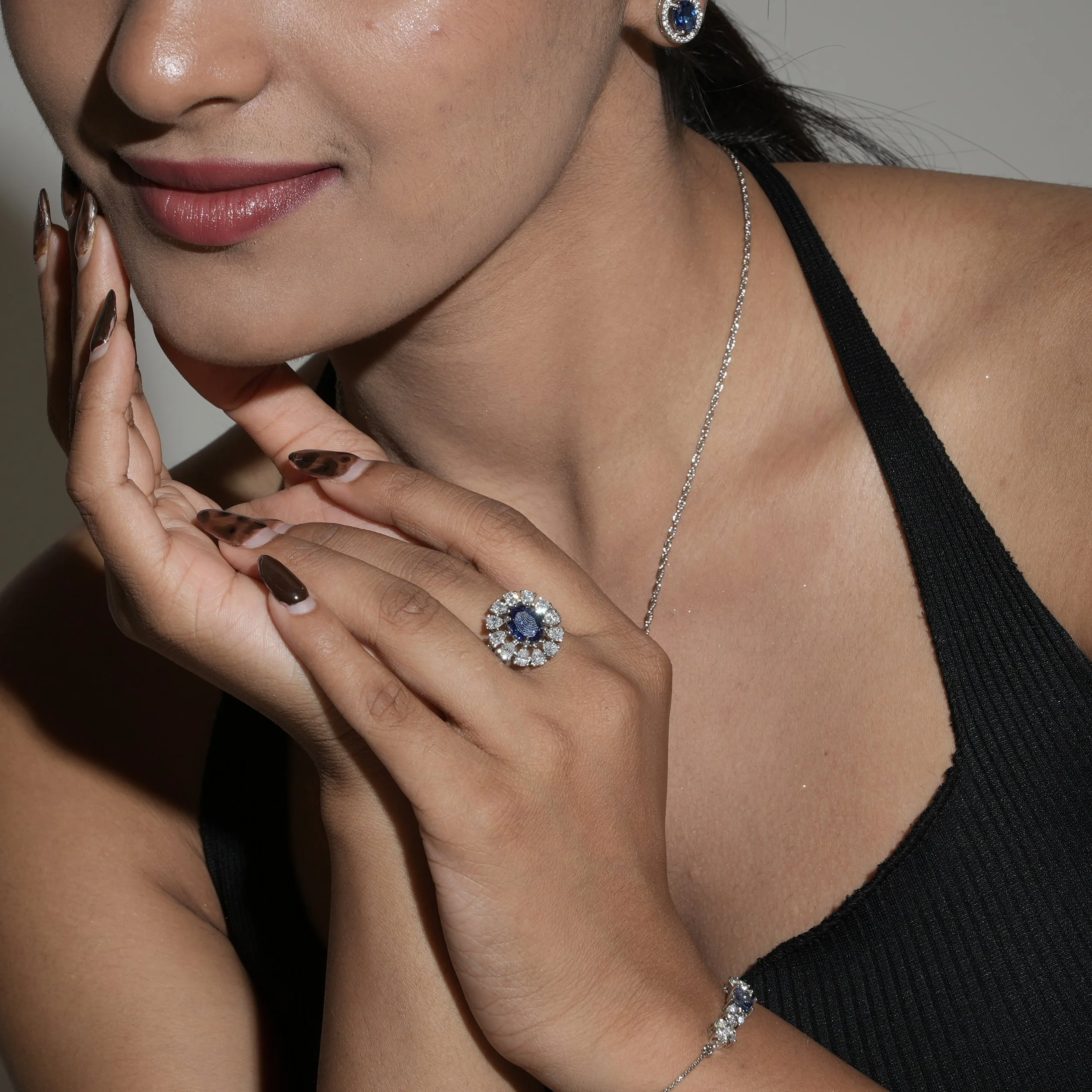 Close-up of a woman wearing a sparkle necklace, diamond earrings with dark blue center stones, and a black top. She has manicured nails and is posing with her hand near her face, showing a smile and smooth skin.