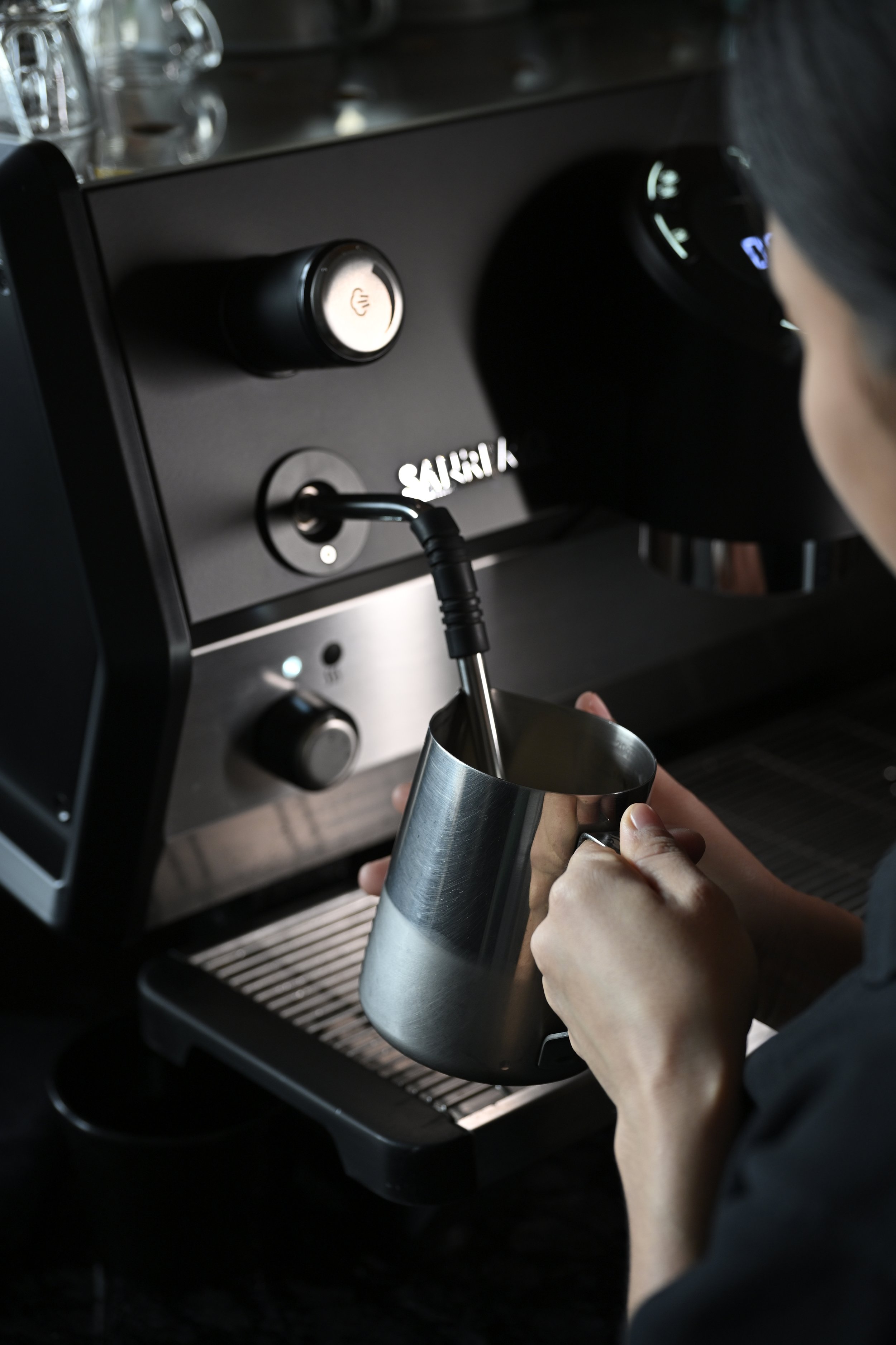 Person holding a stainless steel coffee pitcher under the steam wand of an espresso machine.
