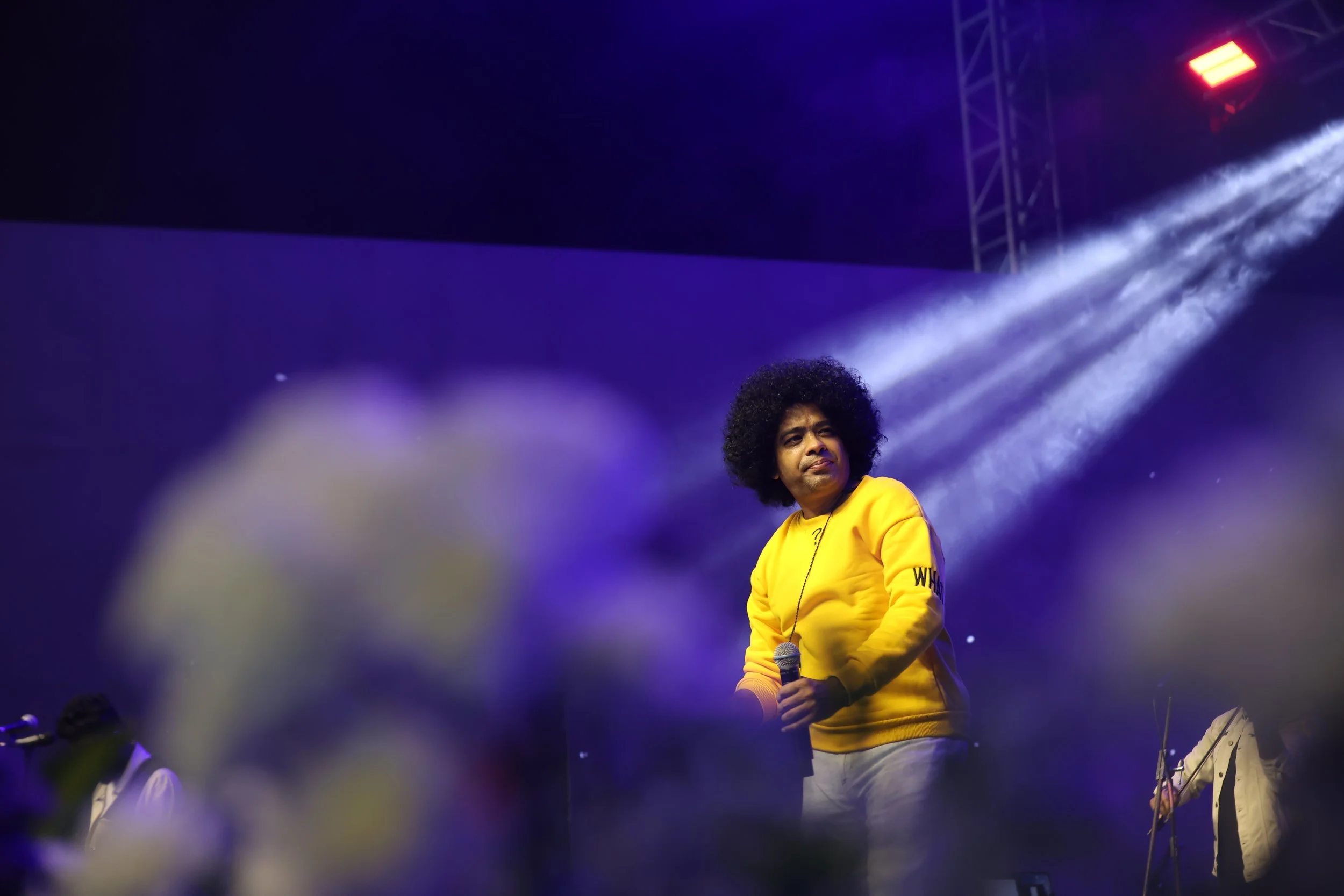 A man with a large afro hairstyle wearing a yellow sweatshirt holding a microphone on a stage with colorful lights and fog effects.
