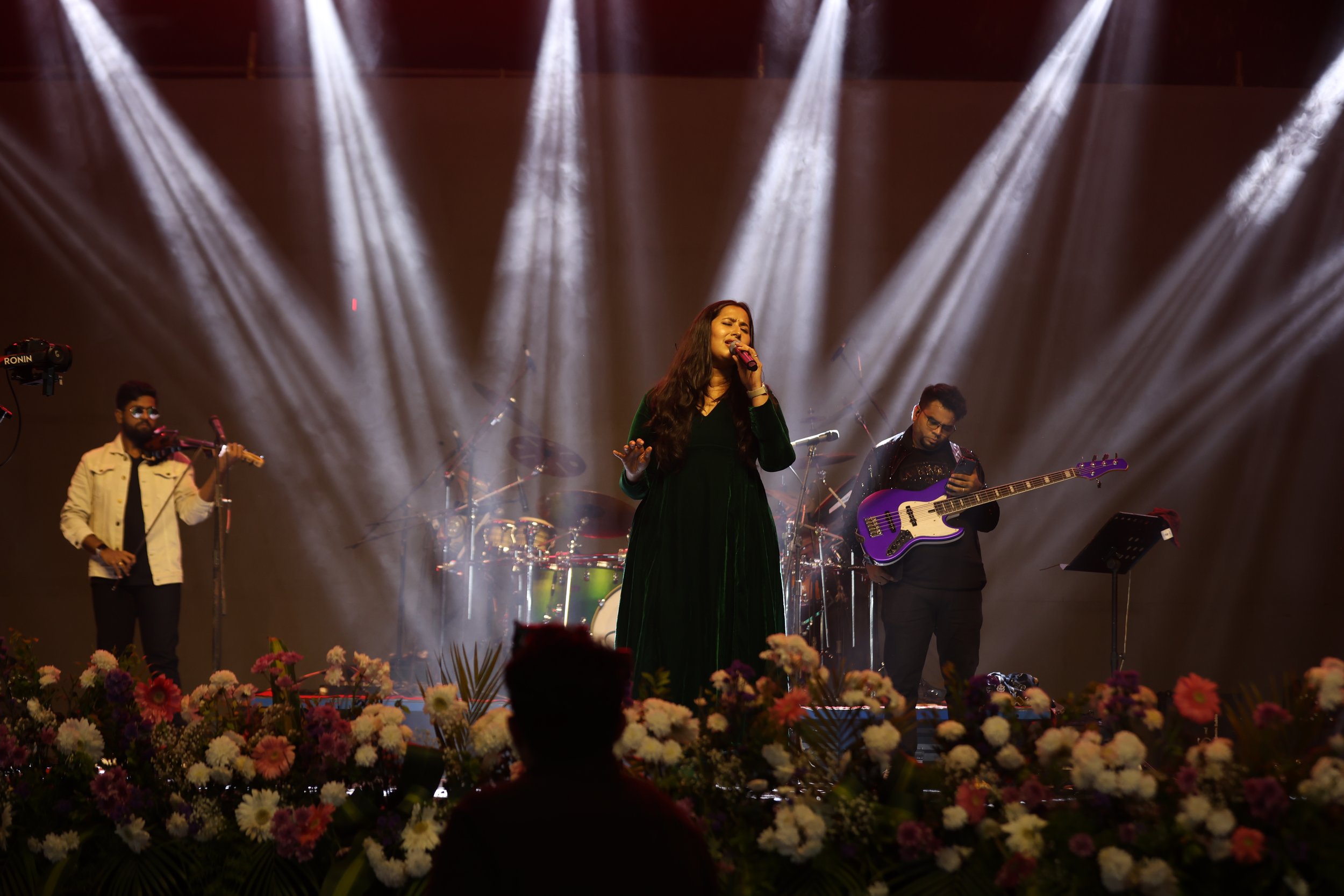 A woman singing on stage with a microphone, surrounded by a band including a guitarist and drummer, with stage lights shining down and flowers at the front of the stage.