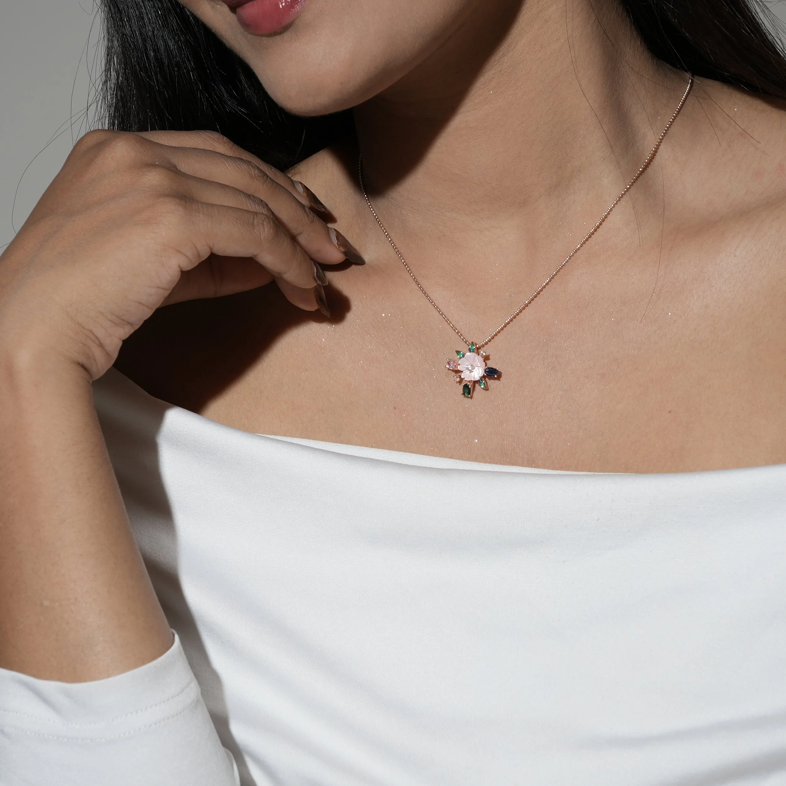 Close-up of a woman wearing a necklace with a floral pendant and a white top. Her hand is near her neck, displaying manicured nails with a leopard print design. Her dark hair is visible, and her lips are slightly parted.