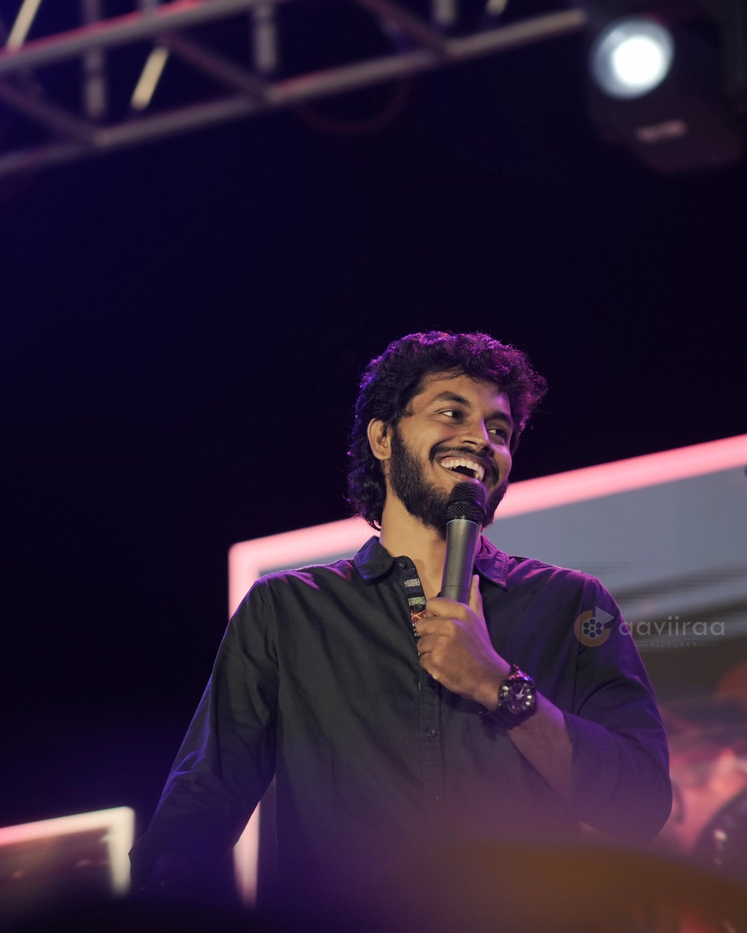 A man with dark curly hair and a beard, smiling and holding a microphone, stands on a stage with purple lighting. The background shows some stage equipment and a pinkish horizontal line.