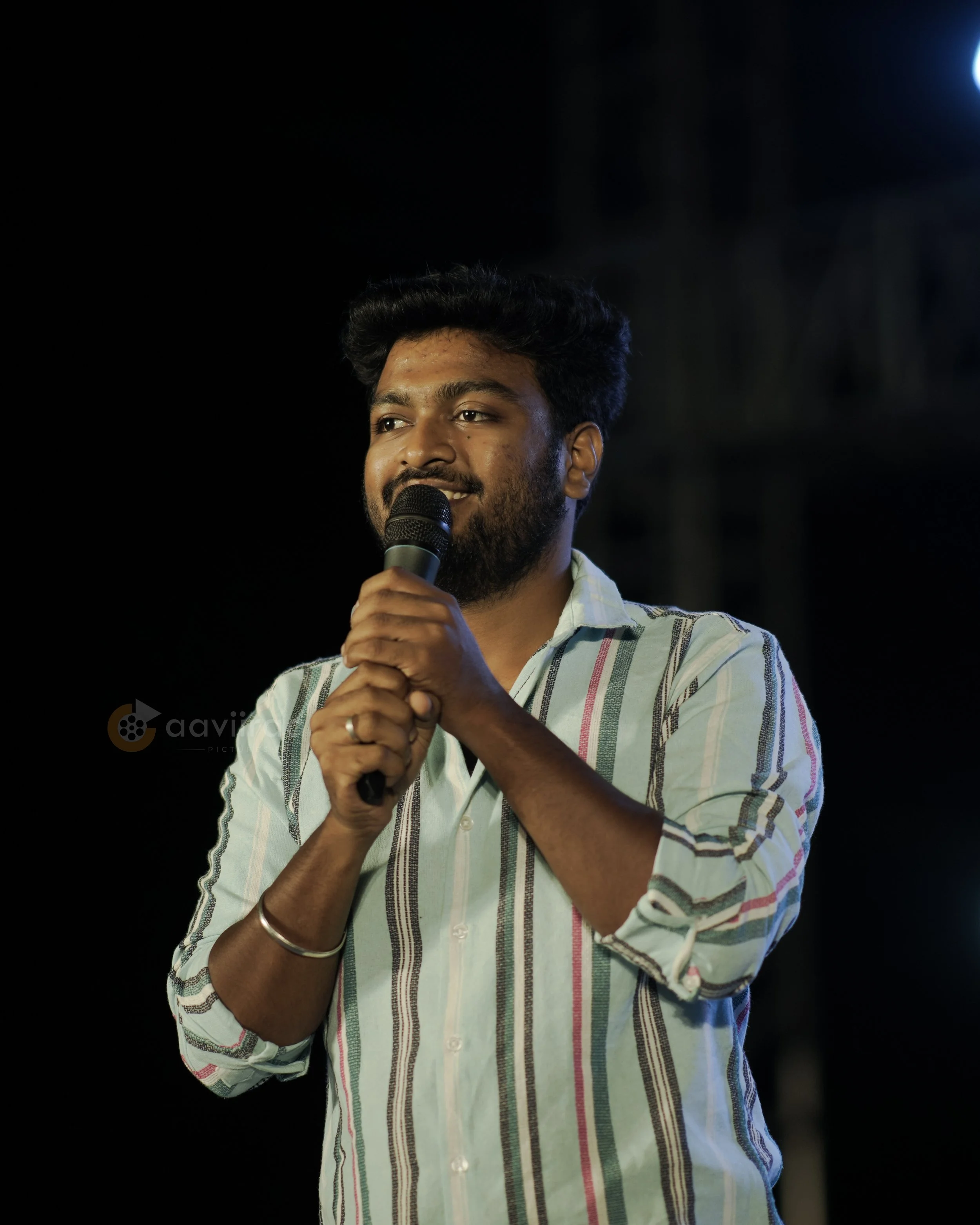 A man with dark hair and beard holding a microphone, wearing a striped shirt, standing against a dark background, possibly performing or speaking.