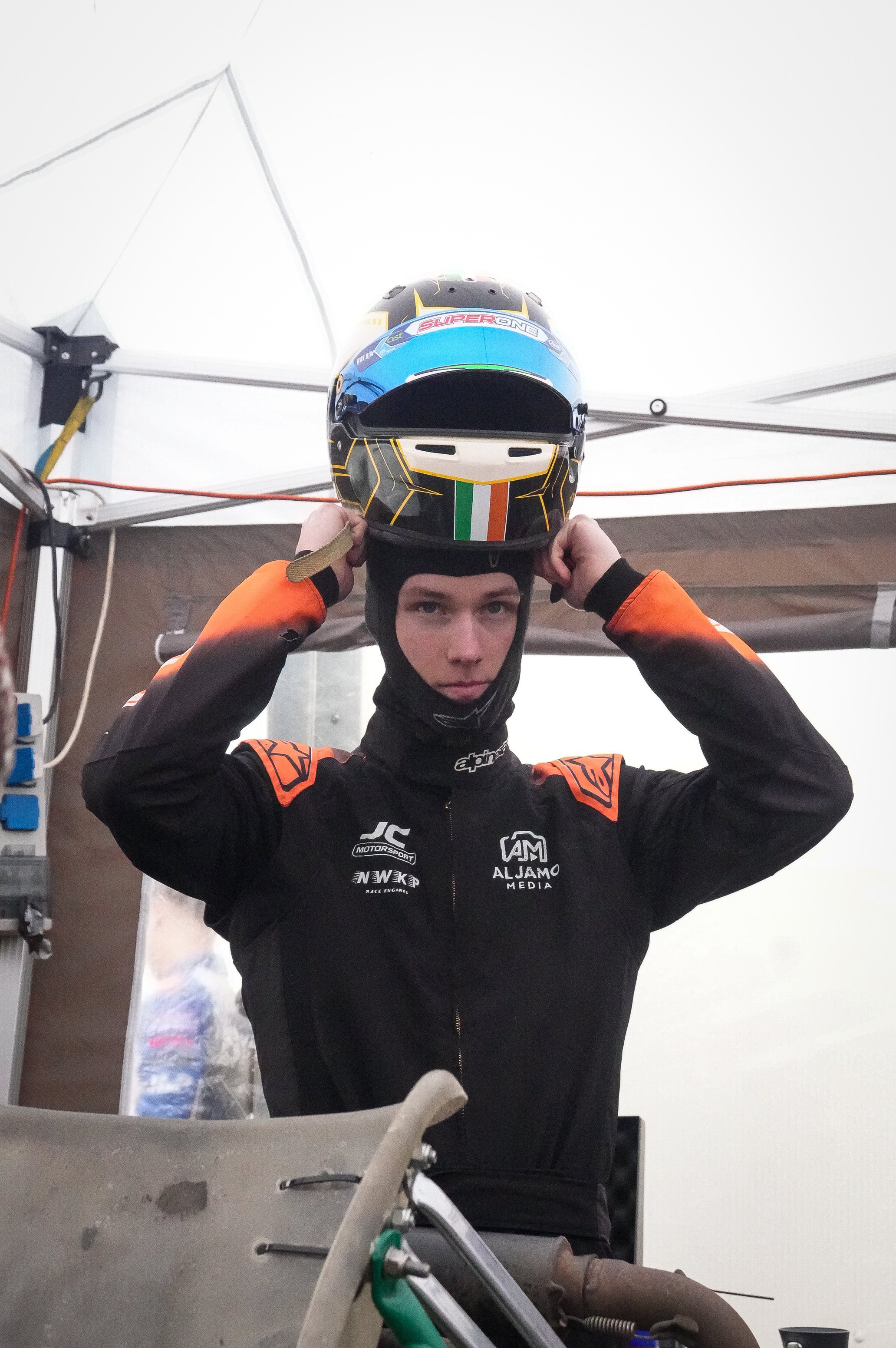 A person in racing gear adjusting a helmet inside a garage or pit area.