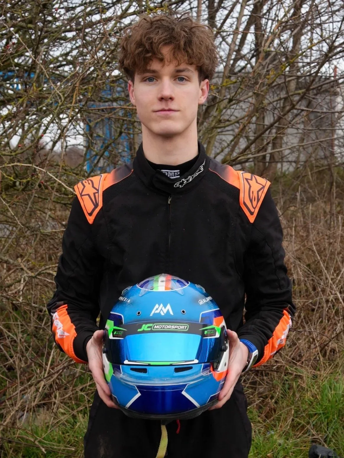 Young man in racing suit holding a blue, green, white, and black racing helmet, standing outdoors in front of leafless trees.