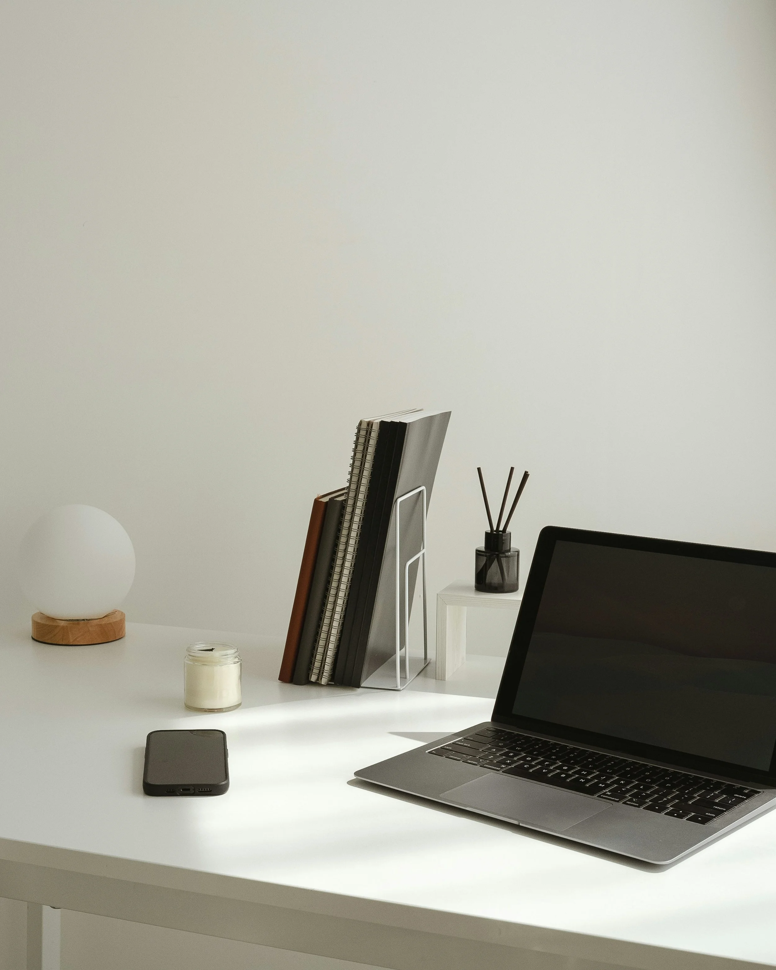 A white desk with a black laptop, a smartphone, a white candle in a jar, a lamp with a round white shade, and a stack of notebooks and files on a white wire rack. There is a white nightstand with a black vase holding reed diffusers.