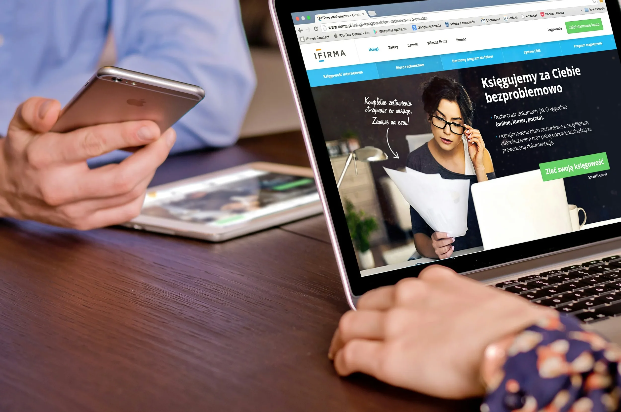 Person holding an iPhone while browsing a website on a MacBook laptop with a Polish language accounting service, in a workspace setting.