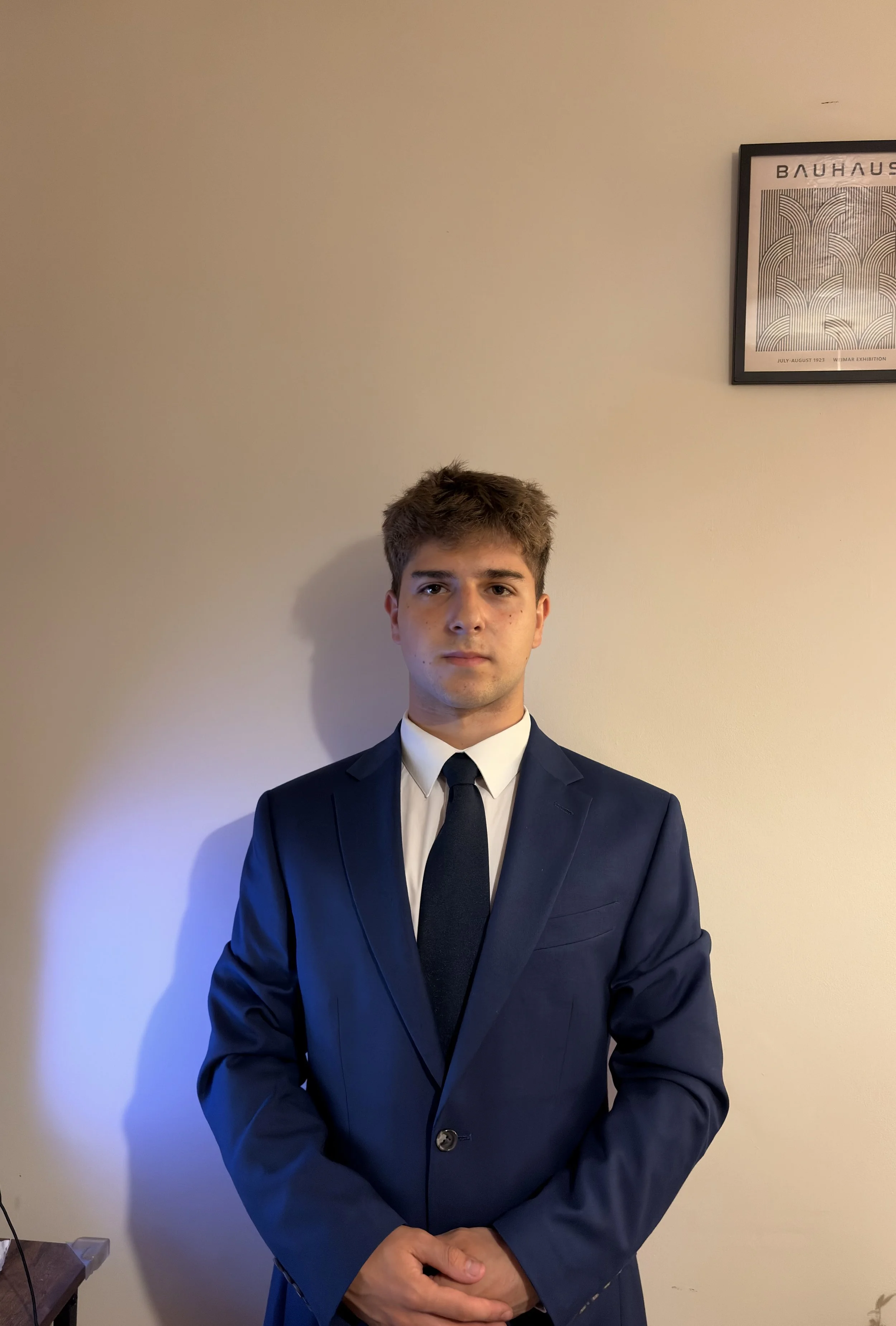 A young man with brown hair and fair skin stands against a beige wall. He is wearing a navy blue suit, white shirt, and black tie, with his hands clasped in front of him. A framed art print with geometric patterns is on the wall behind him.