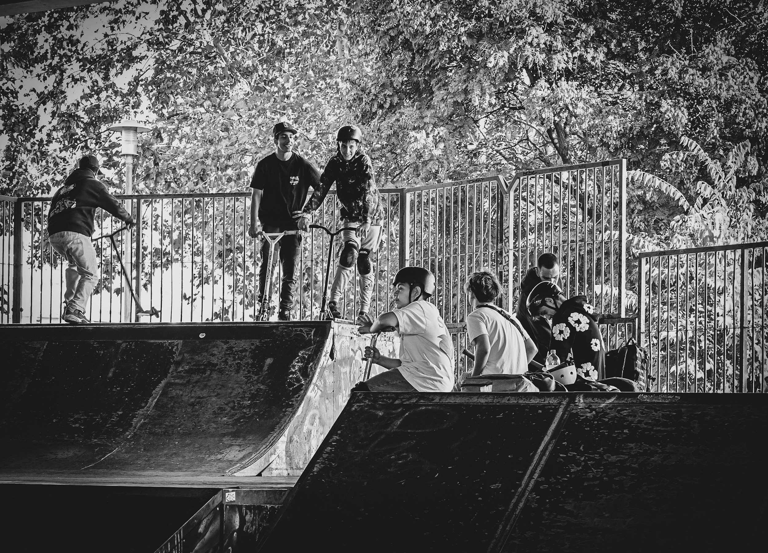 Group of young people at skate park, some on skateboards, some standing or sitting, surrounded by trees, black and white photo.