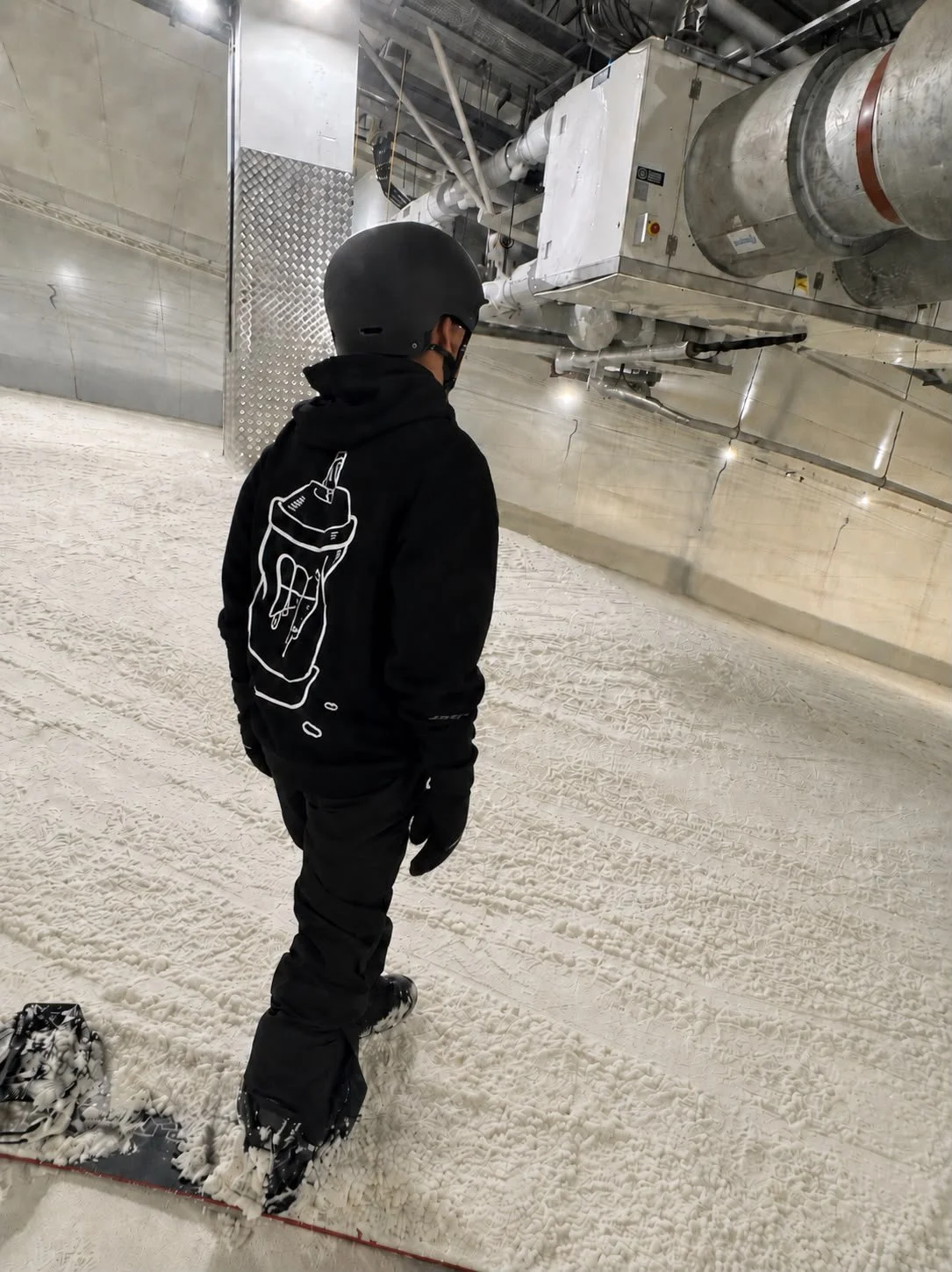 Person wearing a black helmet, black gloves, and a black hoodie with a white outline of a glue bottle on the back, standing on artificial snow inside an industrial or ski facility with large metallic pipes and equipment overhead.