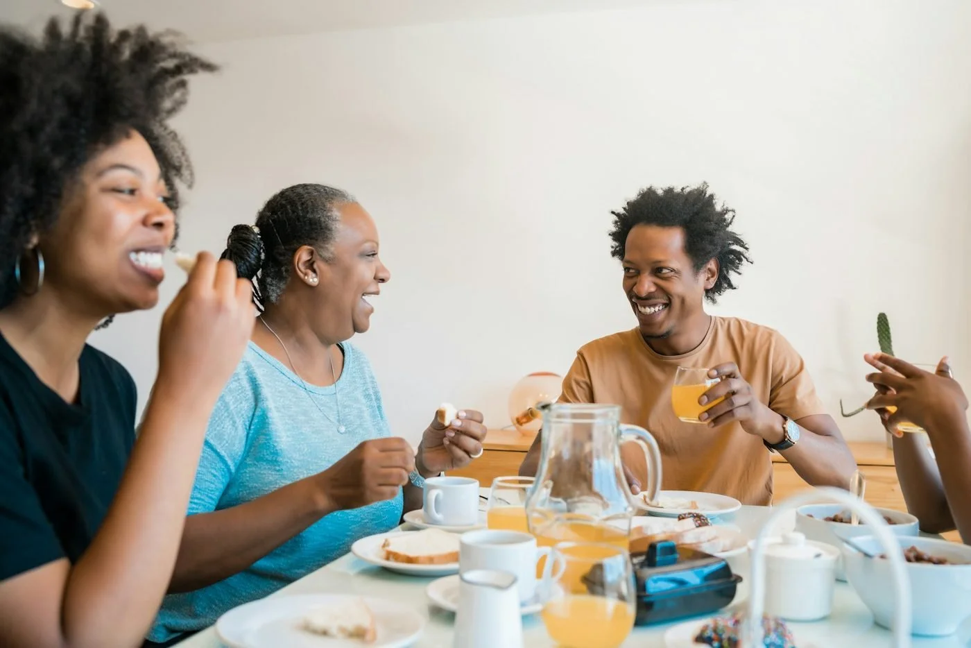 Four friends enjoying breakfast together at a table, smiling and laughing, with plates of food and drinks in a bright, airy room.