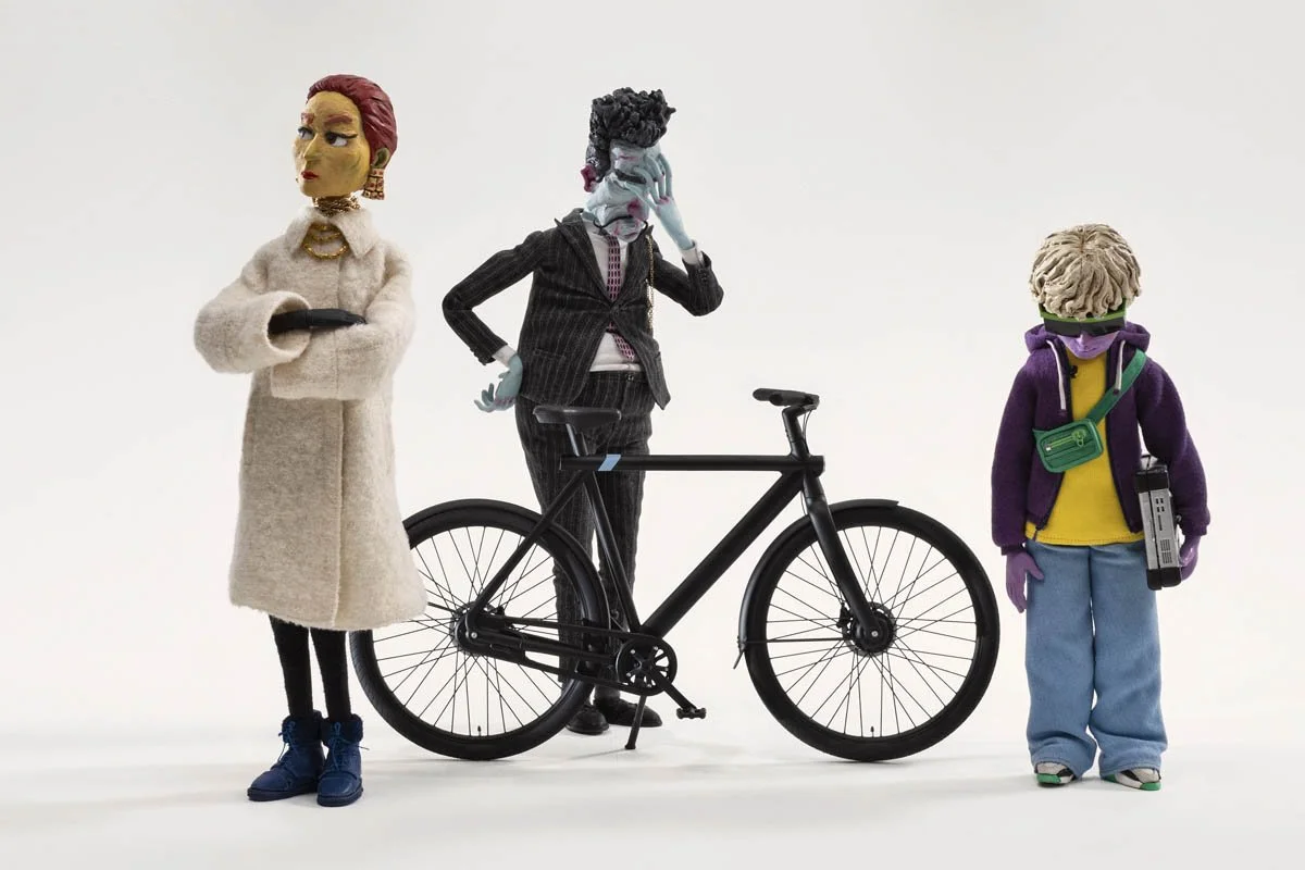 Three dolls with distinct outfits and accessories, standing on a white background, with a bicycle between the dolls.
