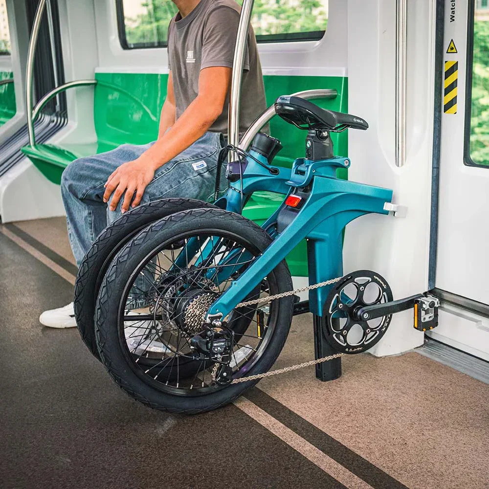 A person sitting on a bench inside a train, with a blue electric bicycle mounted on a bike rack beside them.