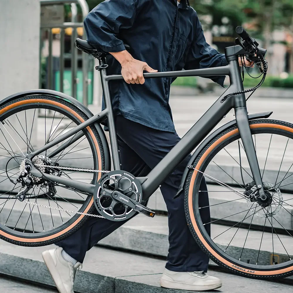 Person holding a matte gray electric bicycle on a city street, wearing a dark jacket and light sneakers.