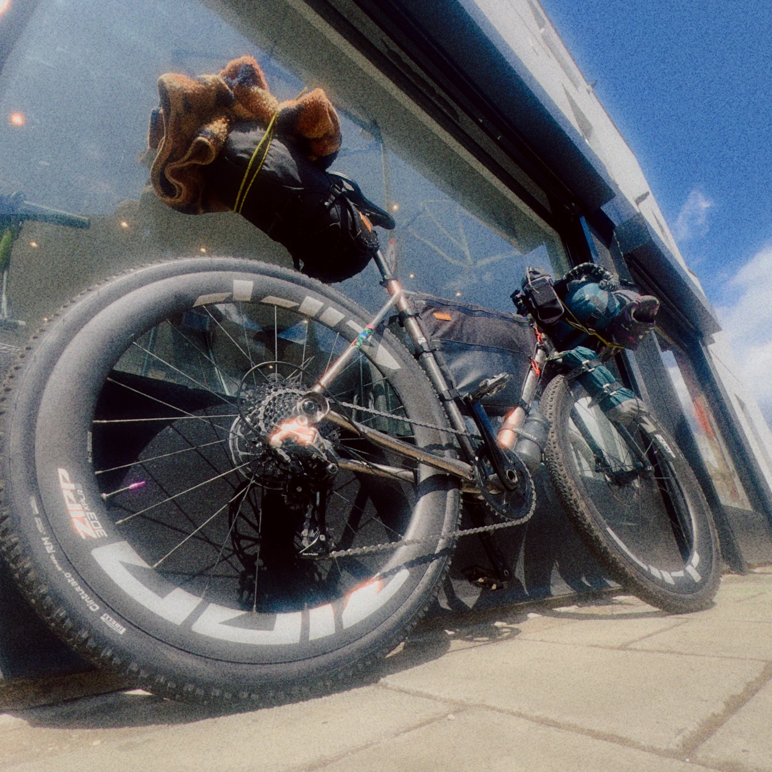 A mountain bike with thick tires and disc brakes parked on a concrete surface behind a glass window, with reflections visible in the glass.