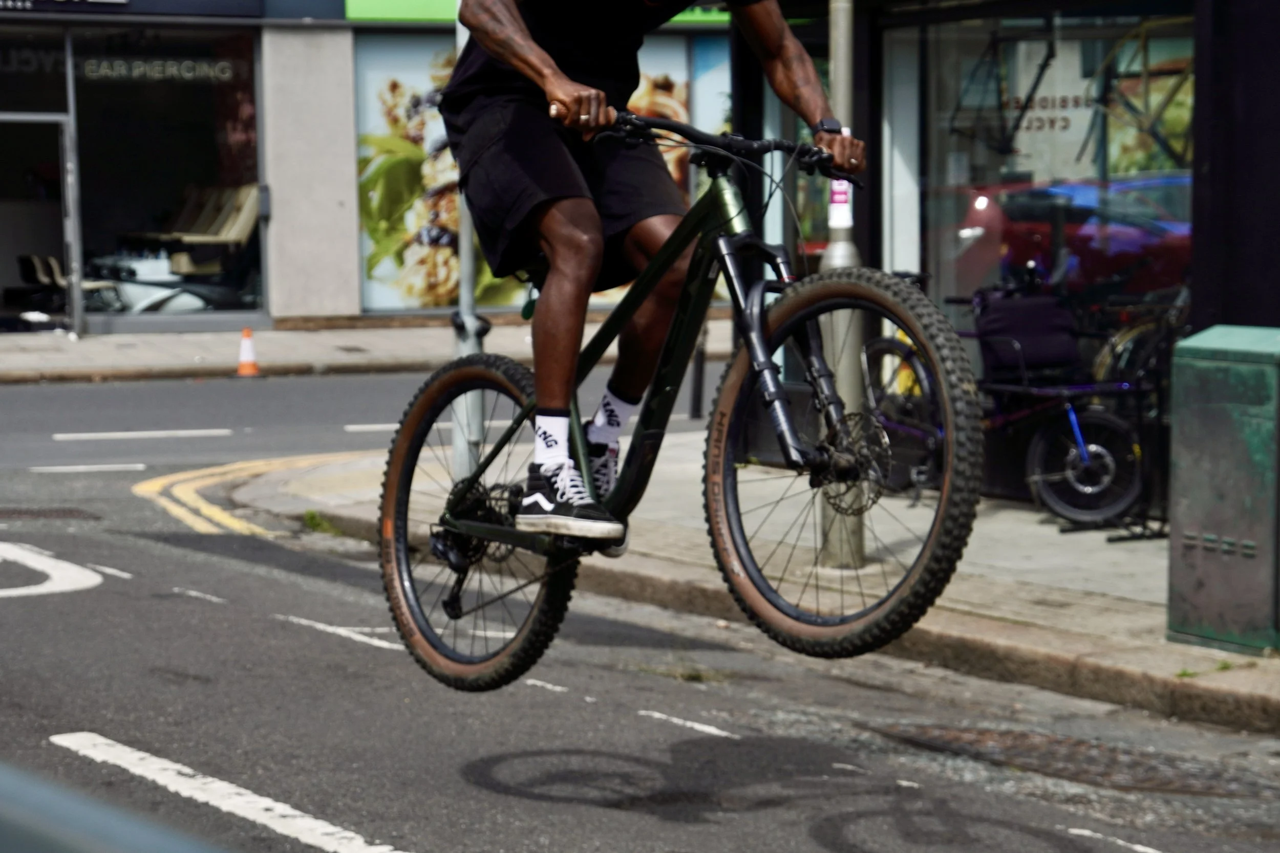 A man riding a mountain bike on a city street sidewalk, wearing black shorts, a black T-shirt, and black sneakers with white soles.