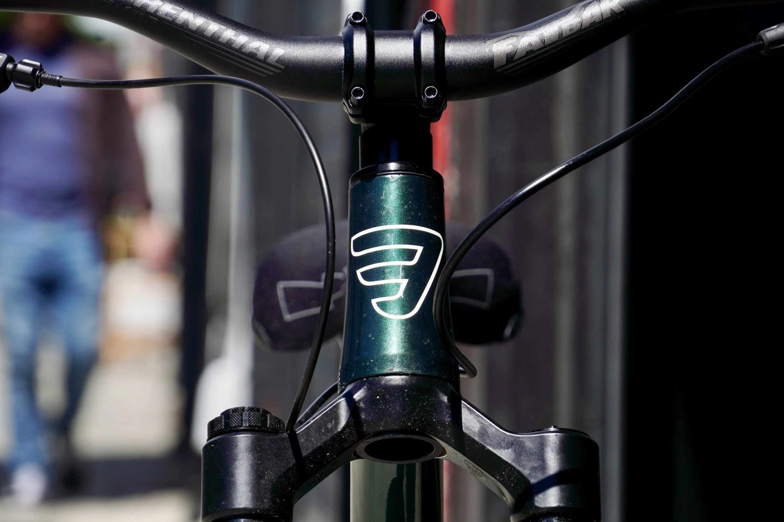 Close-up view of a bicycle's front handlebar and suspension fork, with a blurred background of people and outdoor setting.