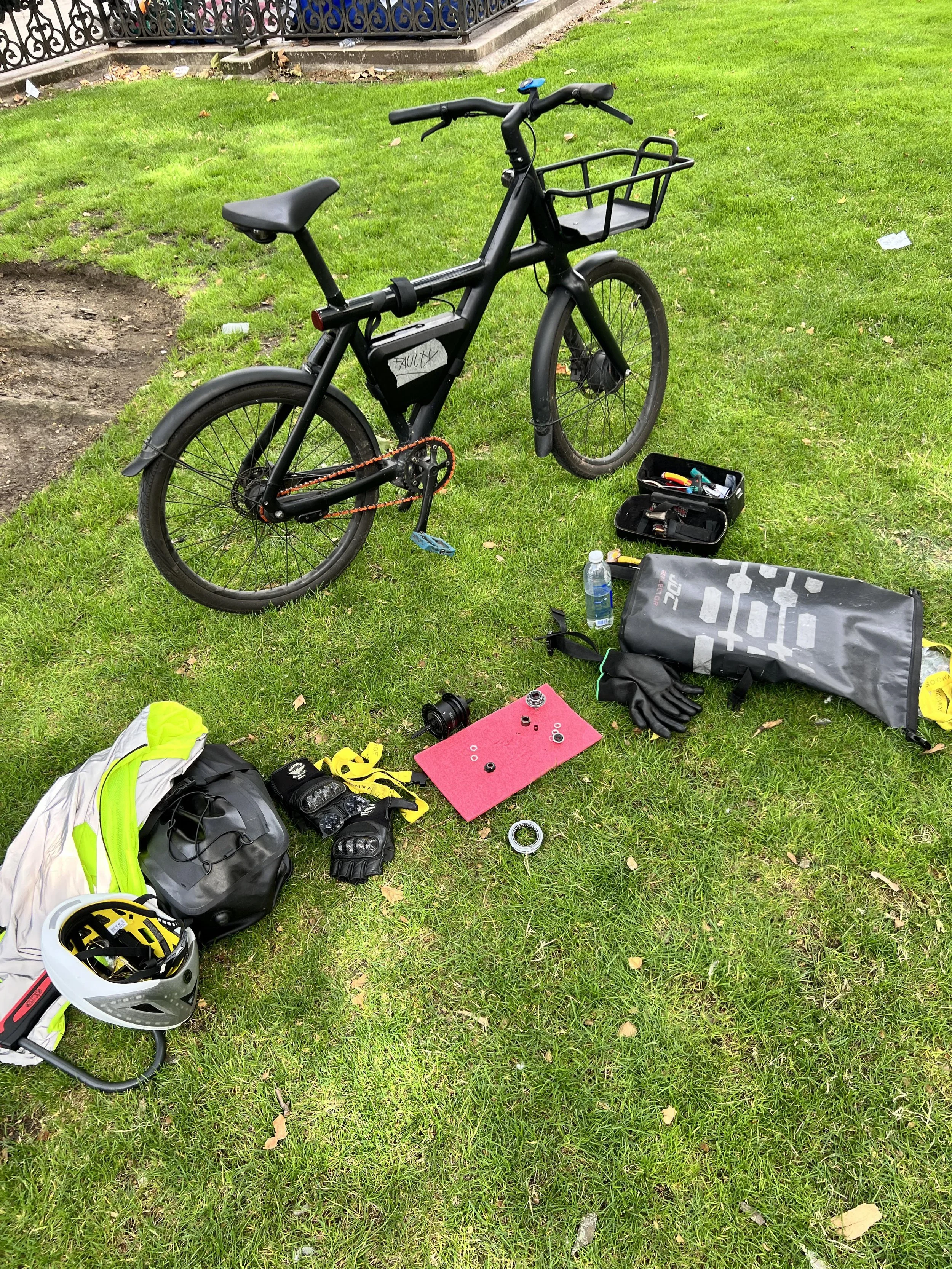 A black electric bicycle with a front basket is parked on green grass. Nearby, there are a white helmet, gloves, a water bottle, a pump, and various tools and gear laid out on the ground, indicating maintenance or repair work.