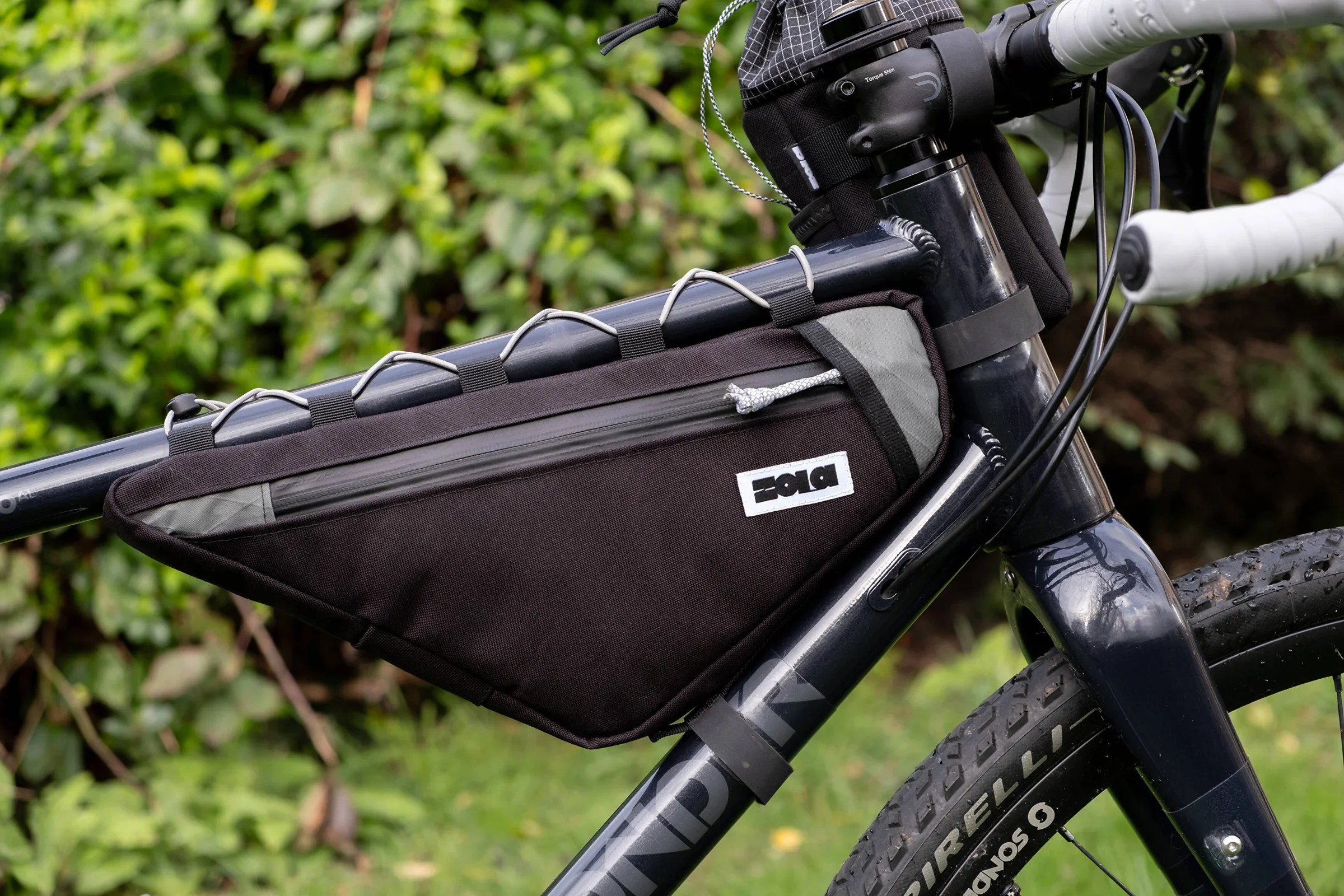 Close-up of a black mountain bike with a black, gray, and white handlebar bag attached to the top tube, with a green, leafy background.