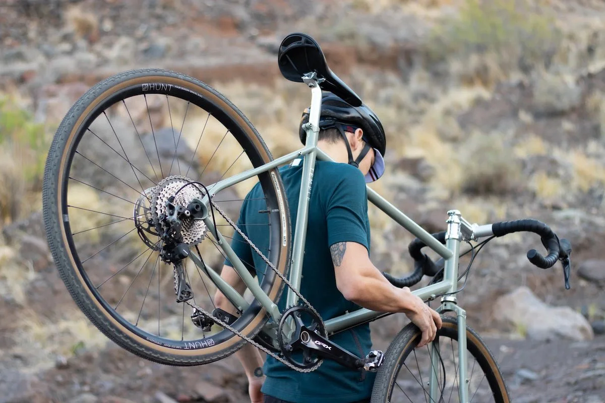 A person wearing a helmet and sunglasses carrying a mountain bike on their shoulder outdoors, with a rocky landscape in the background.
