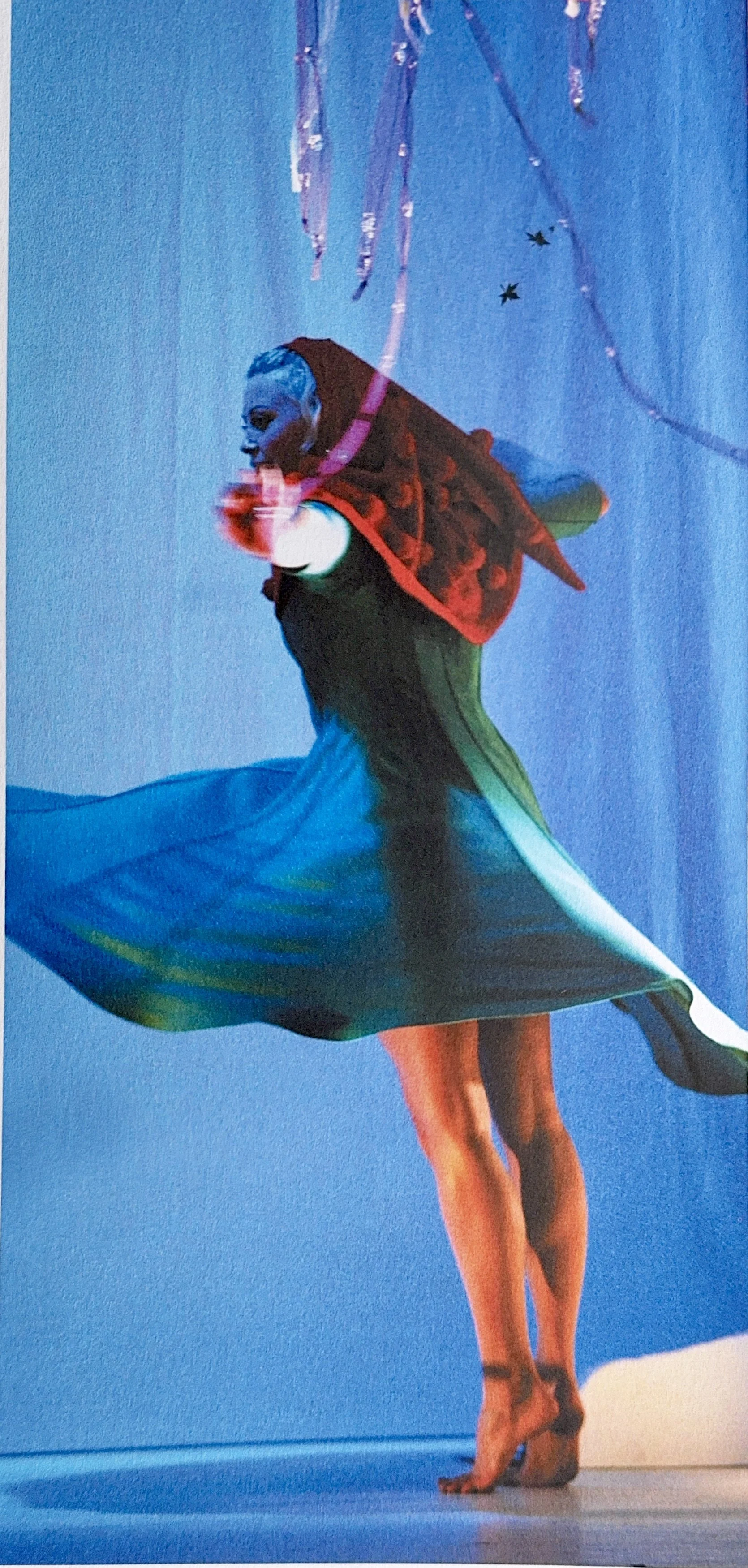 A person wearing a blue mask and a green skirt, appears to be in mid-movement on a stage with blue lighting. They have a red and black patterned garment over their shoulders, with their hair pulled back. The background is blue with some hanging metal