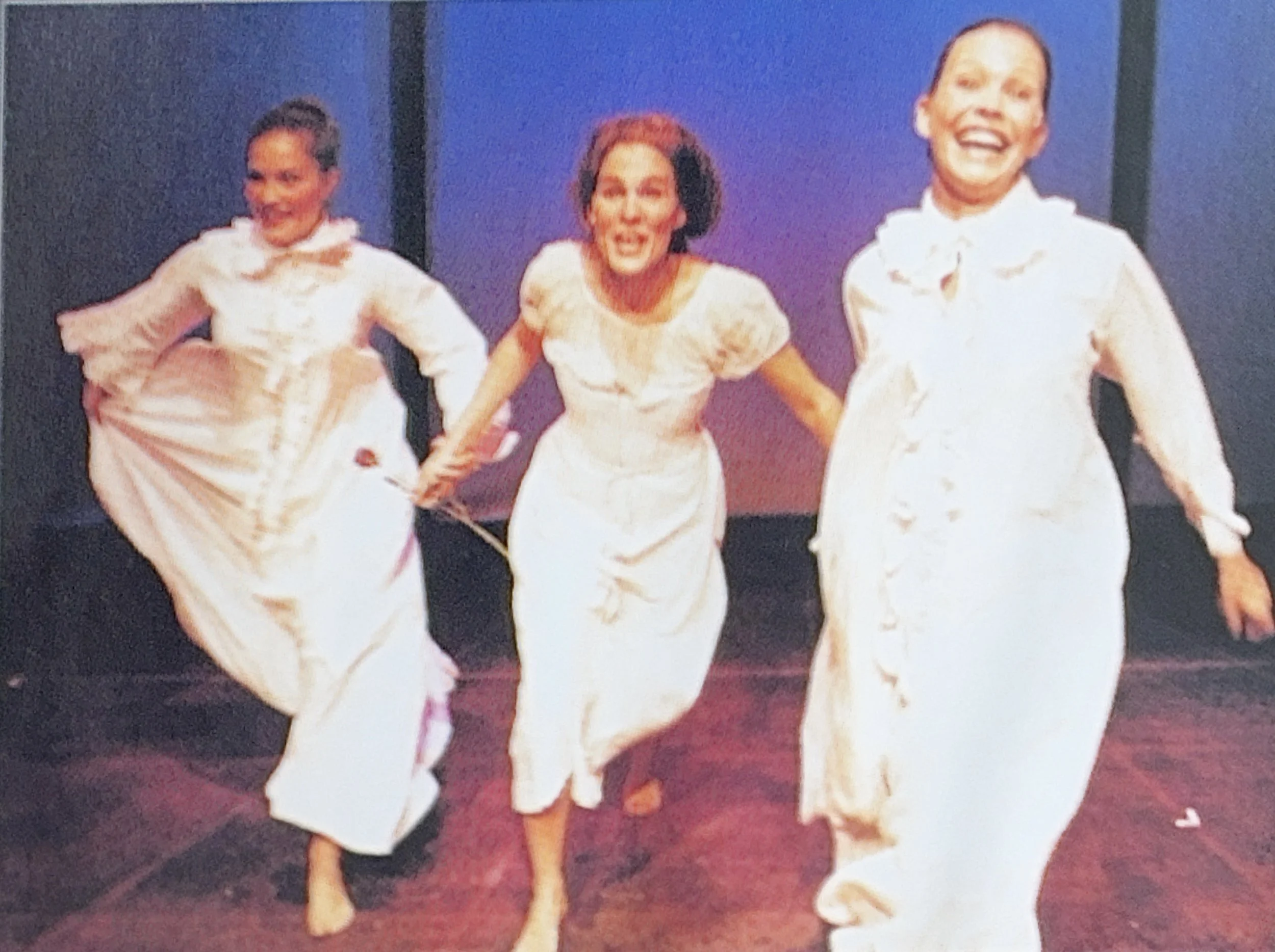 Three women dressed in traditional white religious habits, dancing on a stage with a purple backdrop.