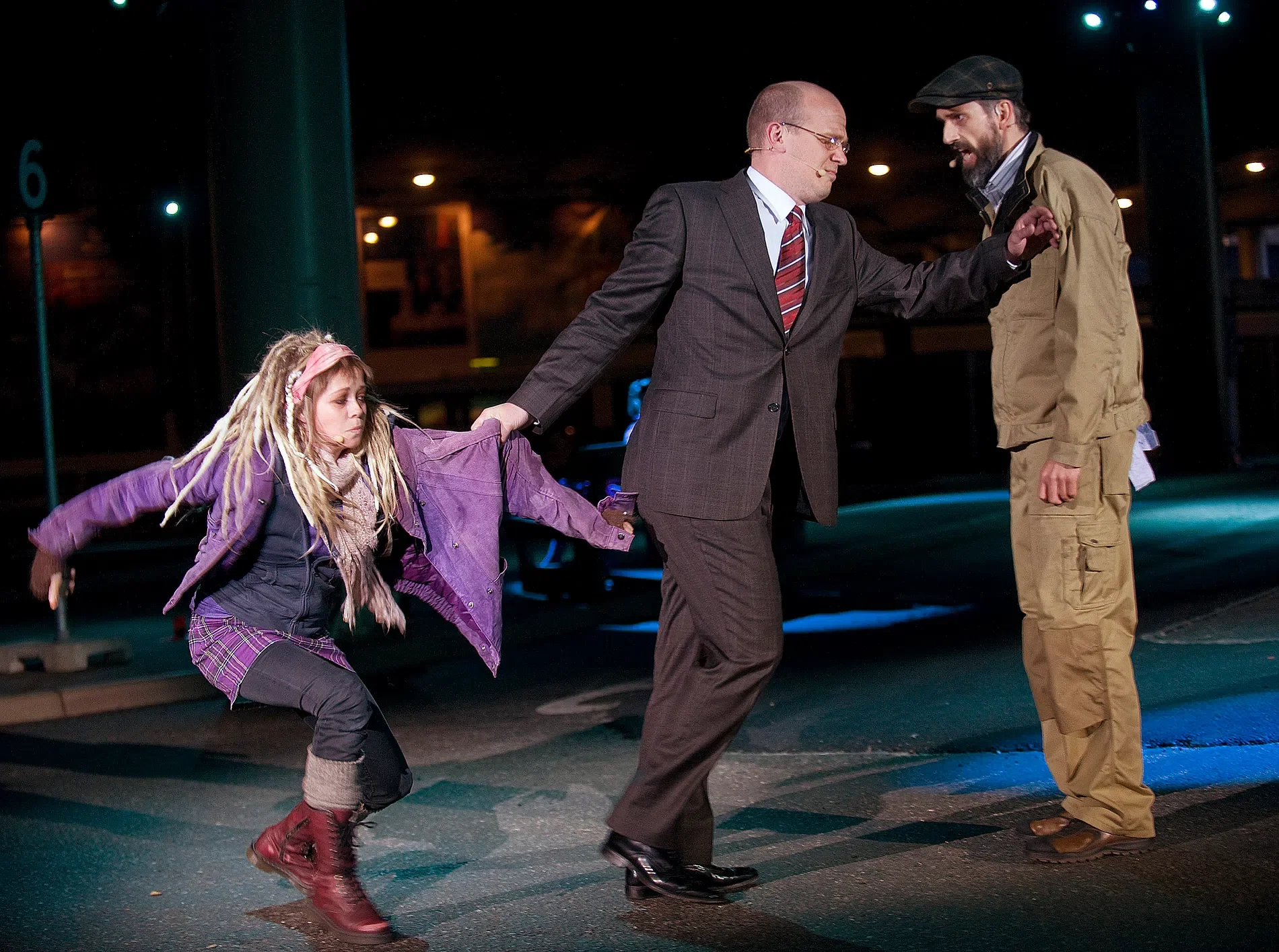 A young girl with dreadlocks, wearing a purple jacket and plaid skirt, pulls on a man's sleeve as he appears to be trying to leave. The girl looks frustrated. The man, dressed in a dark suit, is reaching out to a man in casual clothing, wearing a jacket, tan pants, and a black cap, who is standing with his hand on the man's shoulder. They are on a city street at night.