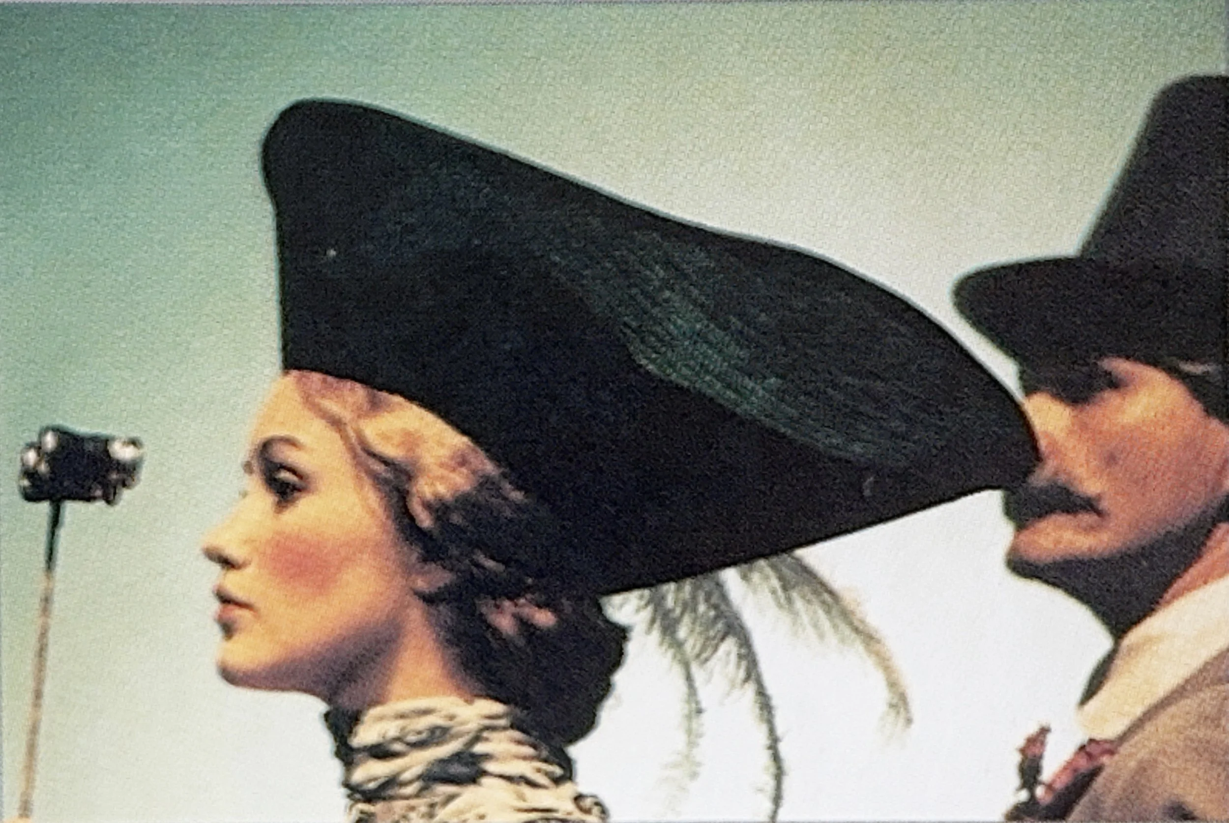 Close-up side view of a woman with makeup and curly hair, wearing a large wide-brimmed black hat. The background shows a clear sky with some streetlights.