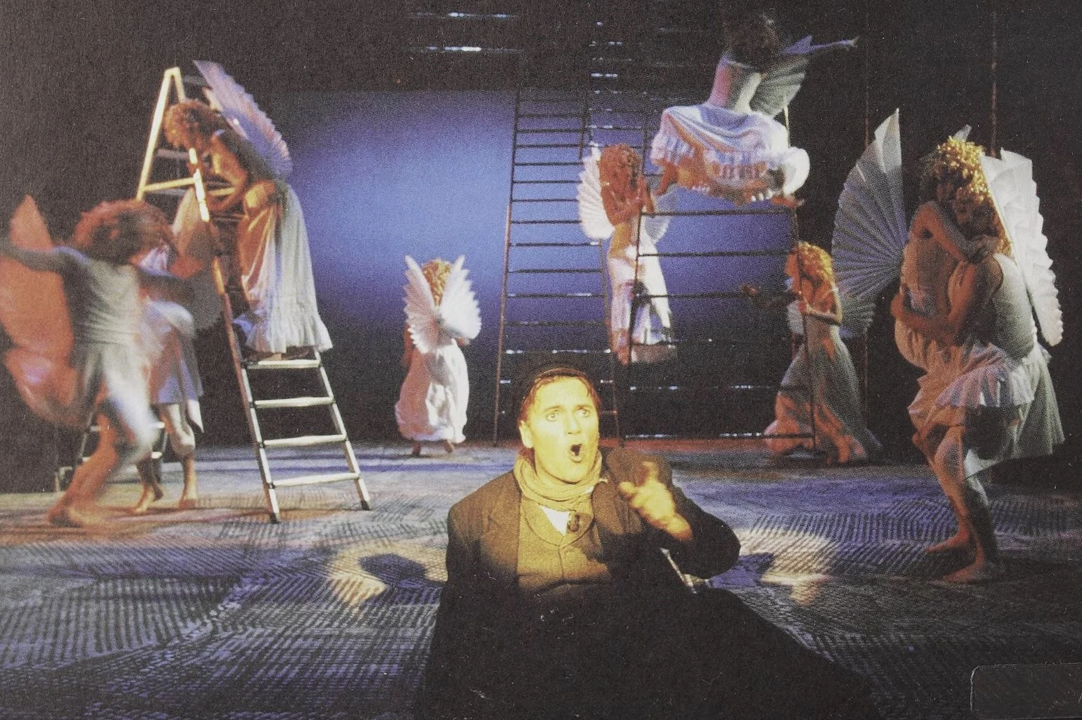 The image shows a theatrical stage scene with several performers dressed as winged creatures and angels, some on ladders and others standing or sitting on the floor. A man in the foreground, dressed in dark clothing, is sitting with an animated facial expression, speaking or singing passionately.