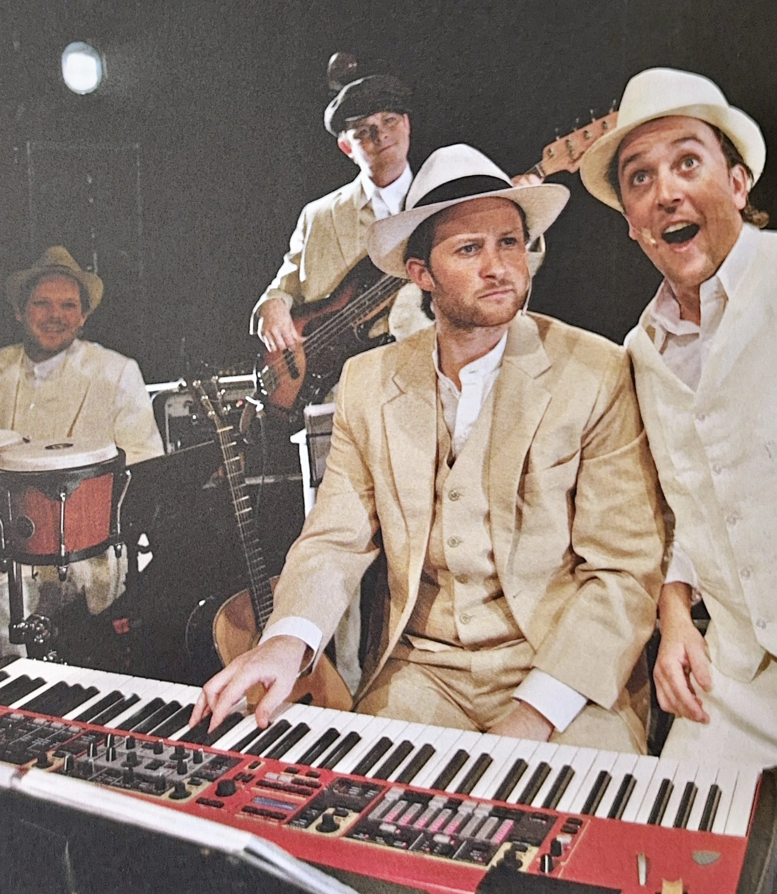 Group of men in vintage suits and hats playing musical instruments, including a keyboard, guitar, and drums, in a studio or performance setting.