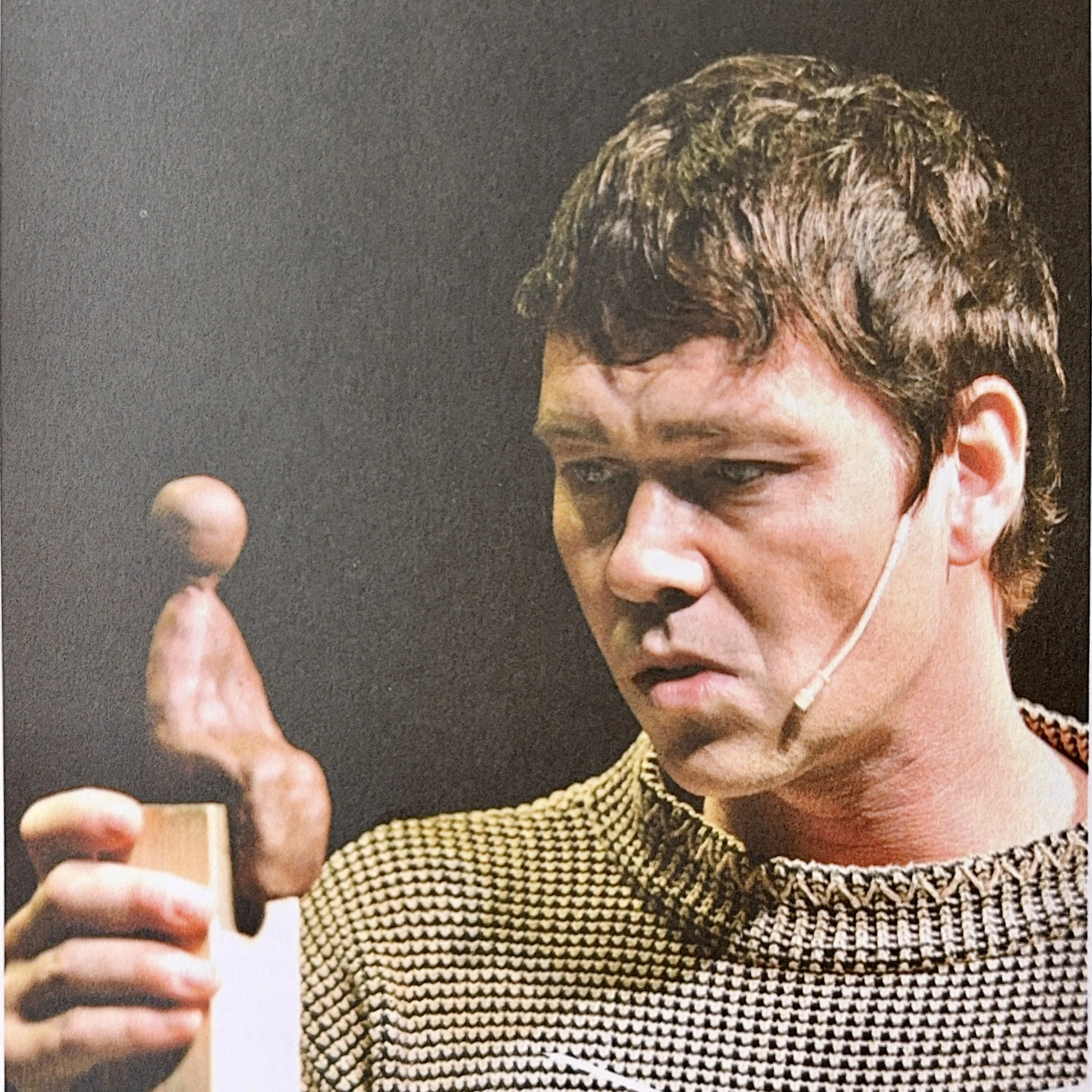 A young man with brown hair taking a selfie with a smartphone, wearing a black and beige patterned shirt and a face mask hanging off his ear.
