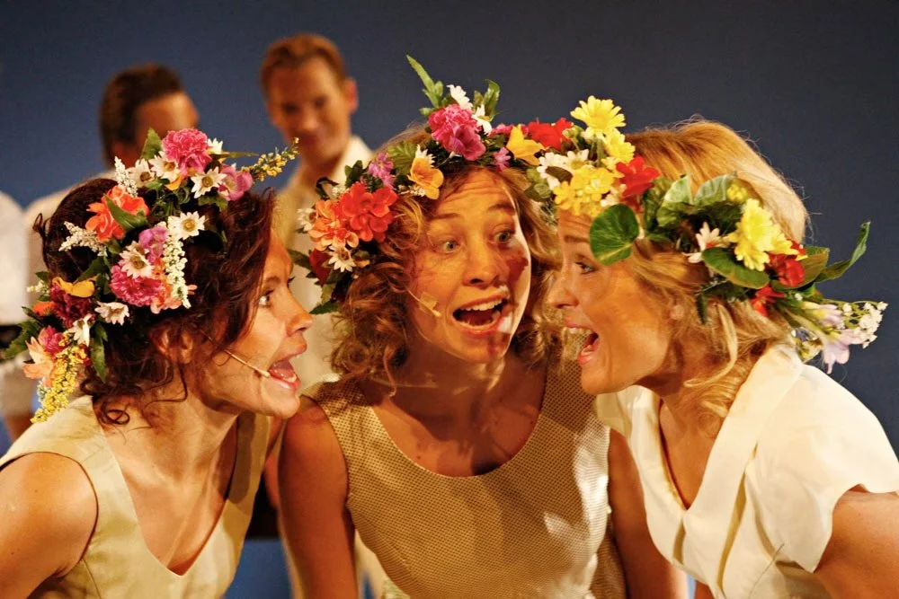 Three women wearing flower crowns are happily talking close together at an event, with two more women smiling in the background.