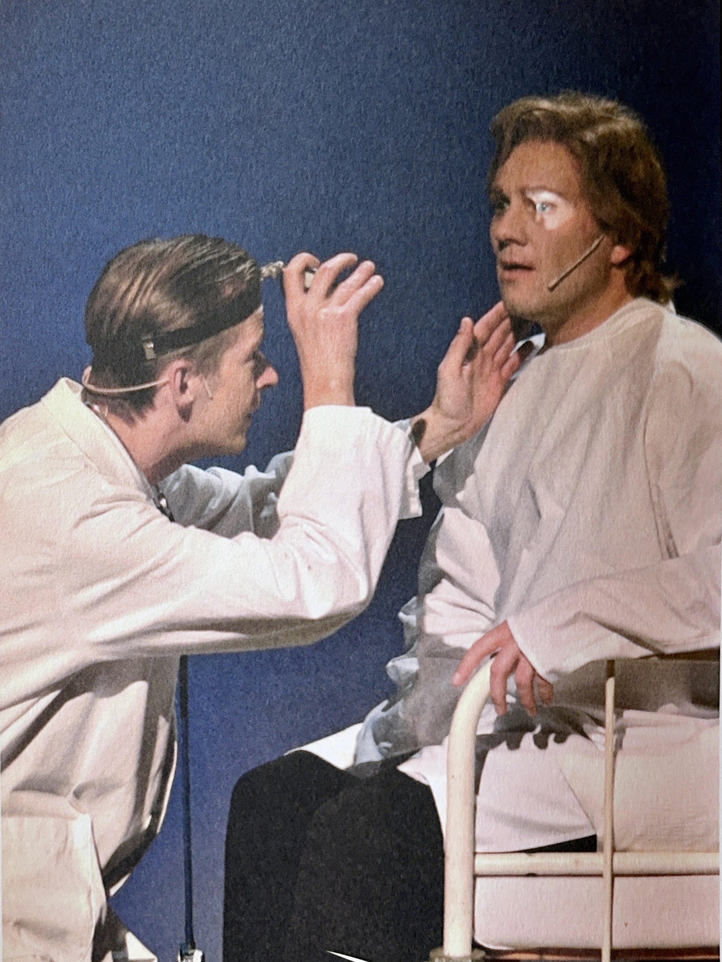 Two men in white coats, one with a headset microphone, seated facing each other, engaging in a medical or consultation session. One man is touching the other's shoulder and they appear to be in a clinical setting.
