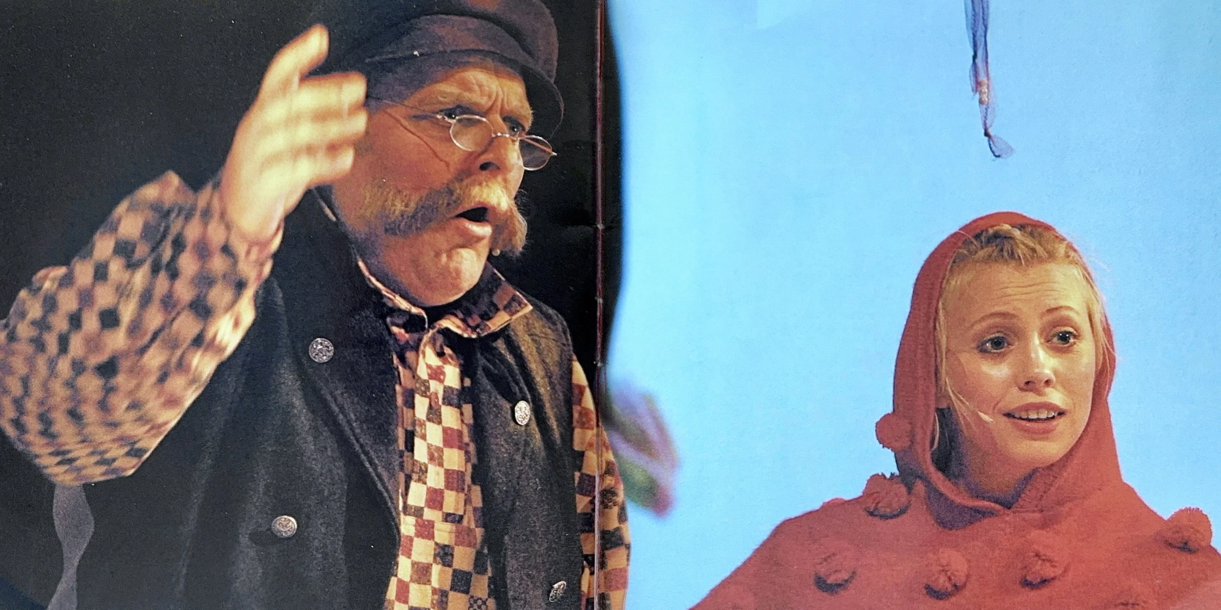 A man with glasses, a beard, and a hat is speaking with an animated expression. He is wearing a patterned shirt and a dark vest. A young woman with red hair, wearing a red hoodie, is listening attentively with a slight smile.
