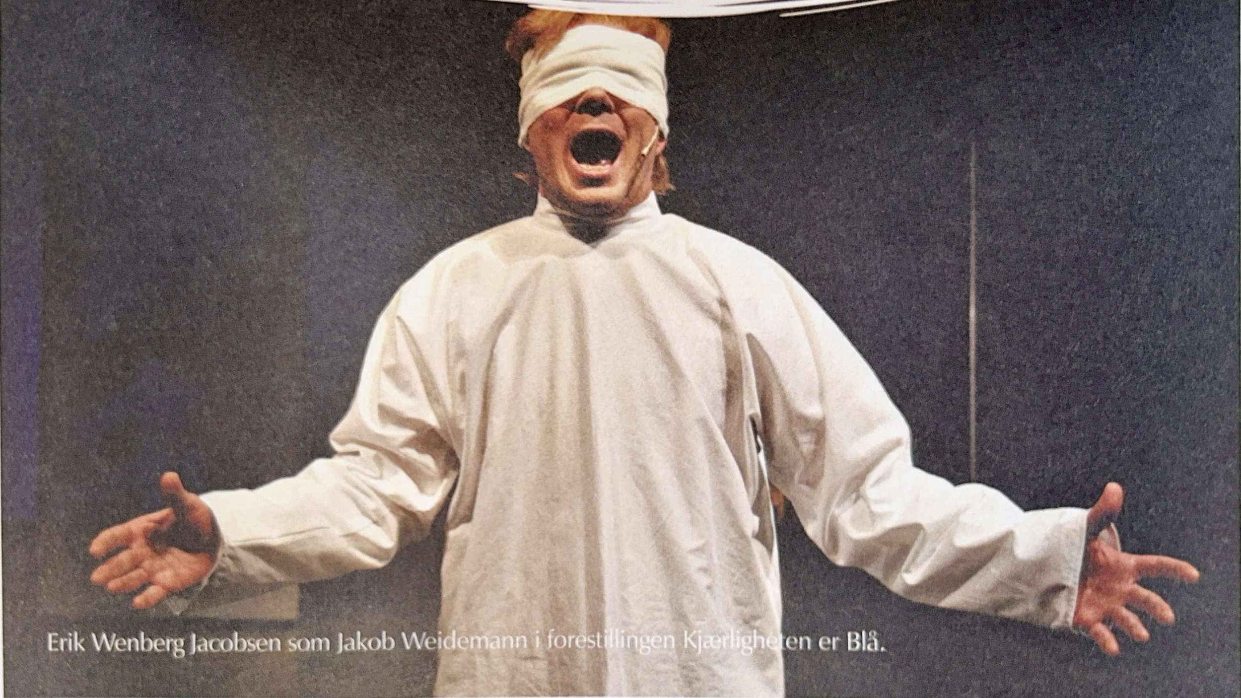 Person dressed in white with a blindfold. Their mouth open as if shouting or singing, and arms extended with palms facing forward. Multiple hands are visible around them, suggesting an audience or group.