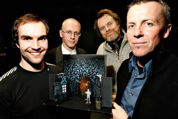 Four men stand around a small, detailed diorama scene in a box, depicting a wooded forest with a house and a miniature astronaut figure inside.