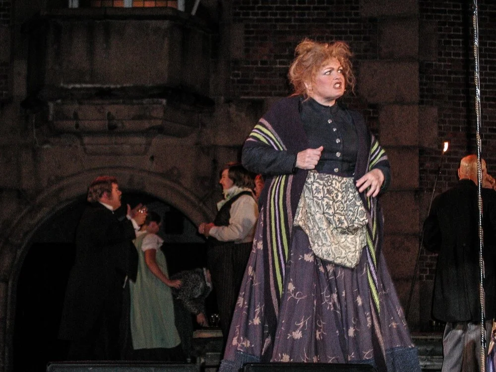 A woman with red hair in period costume on stage, with a distressed expression, in front of a brick building, other actors in period clothing in the background.