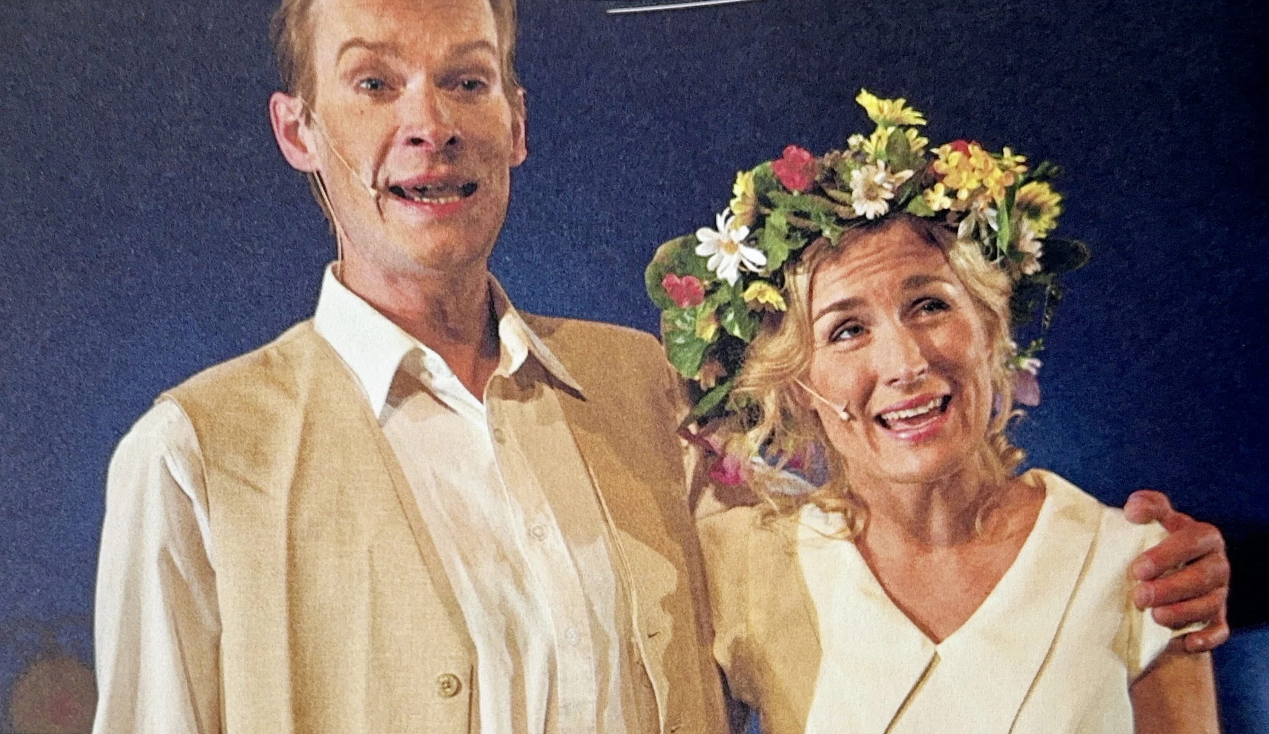 A man and woman on stage, smiling and wearing microphones. The woman is wearing a floral crown and a white dress, while the man is dressed in a beige vest and white shirt.