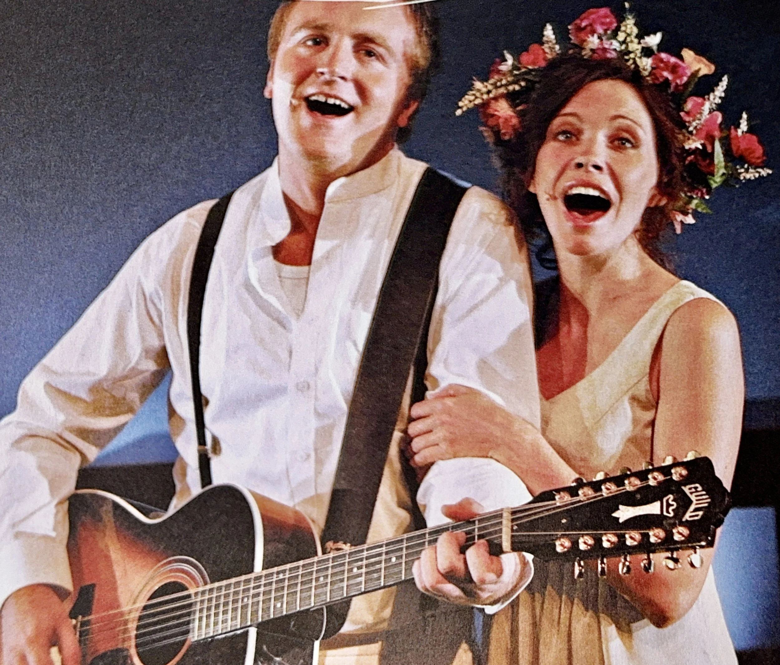 A man playing an acoustic guitar with a woman, both smiling and joyful, in front of a dark background. The woman is wearing a floral headdress.
