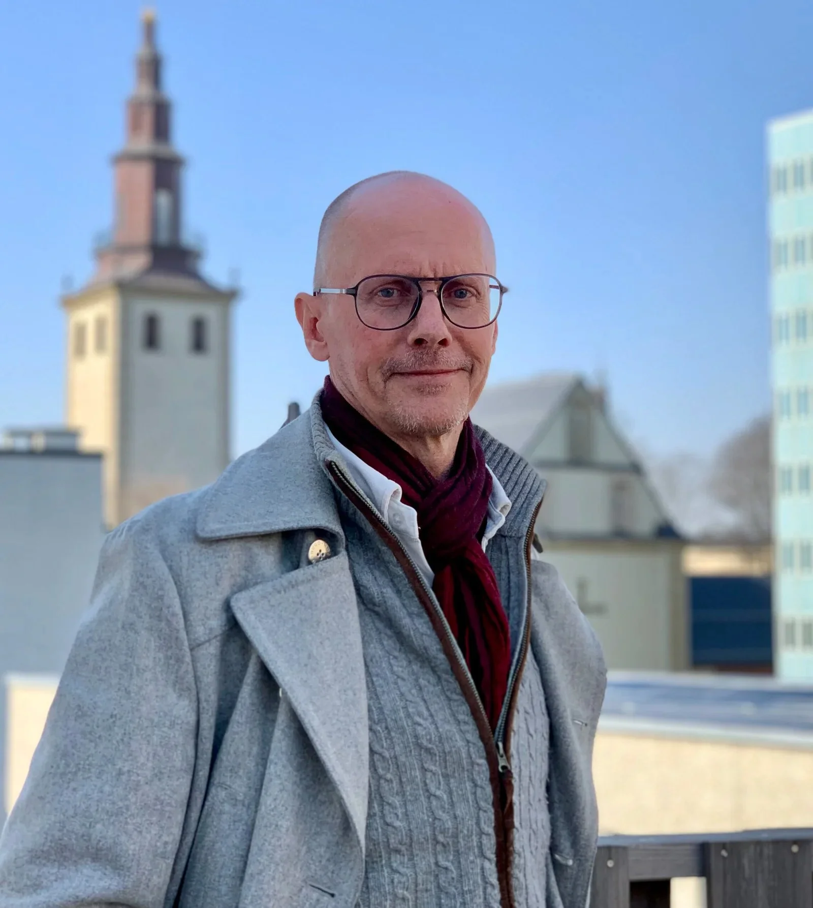 A smiling middle-aged man with glasses, wearing a gray jacket, standing outdoors with a cityscape in the background, including a church steeple and modern buildings.