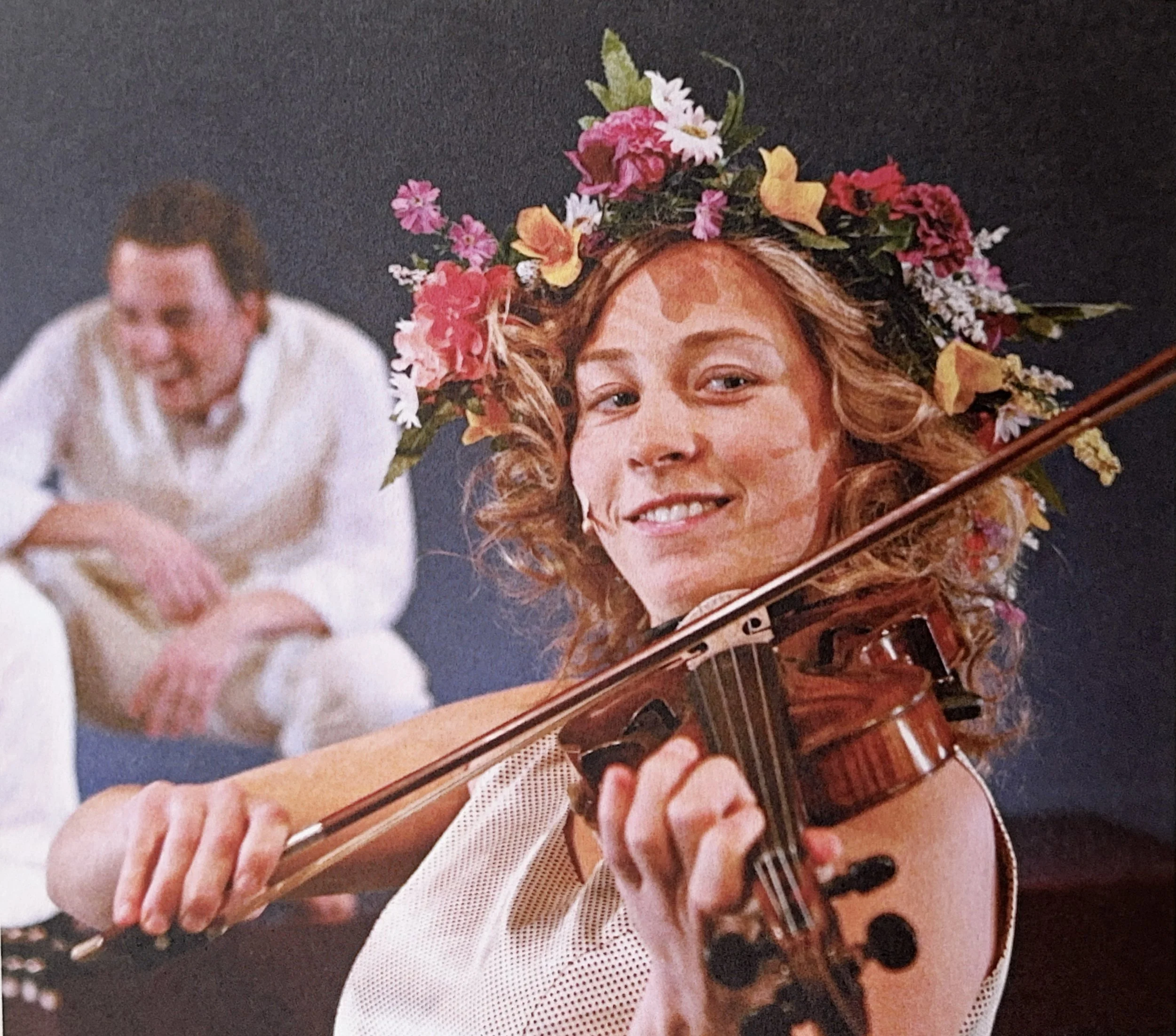 Close-up of a woman with curly hair wearing a floral headband, playing an acoustic guitar, with an out-of-focus man in the background.