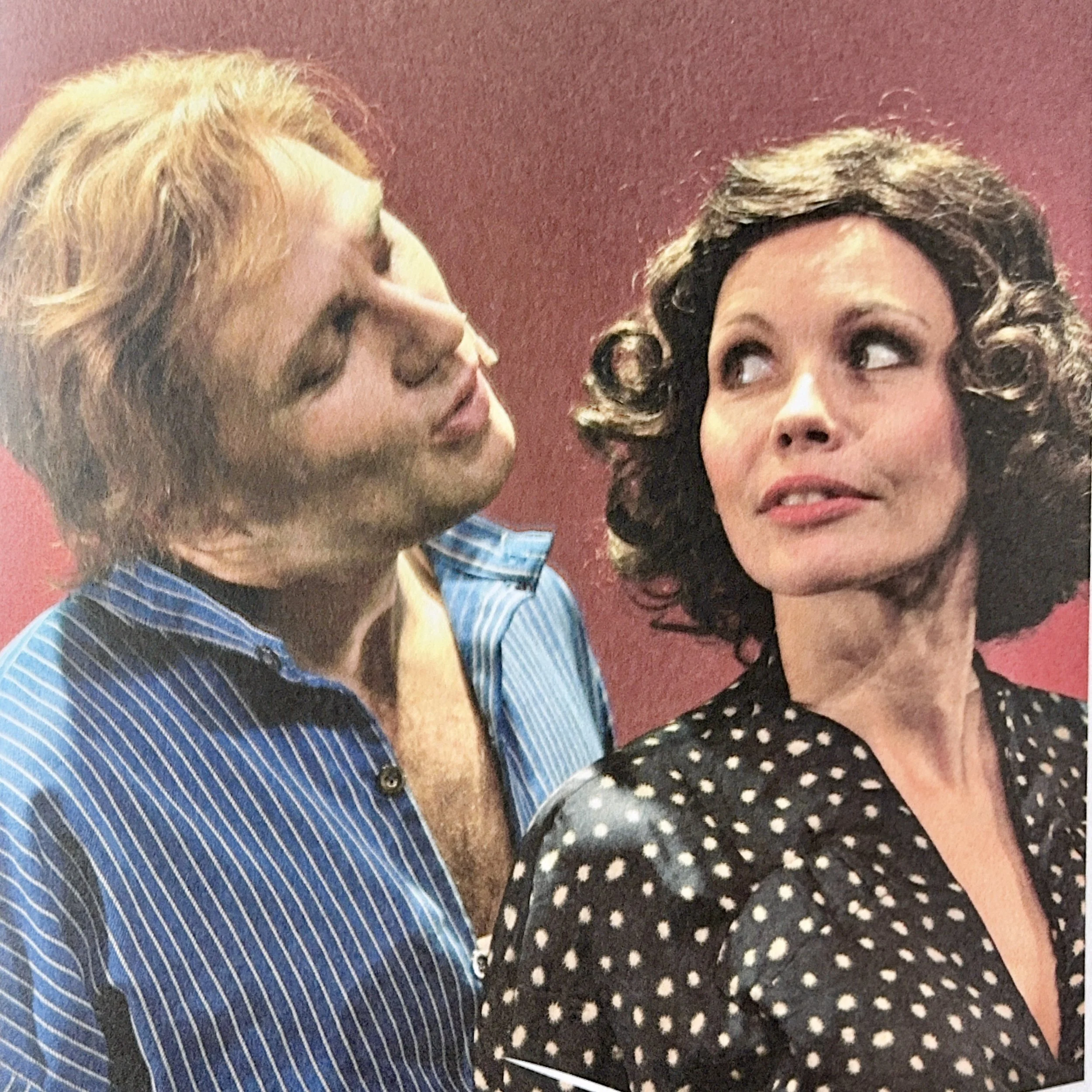 Close-up of two women with vintage hairstyles and makeup, one with light brown hair and the other with dark brown hair with curls, posing against a red background.
