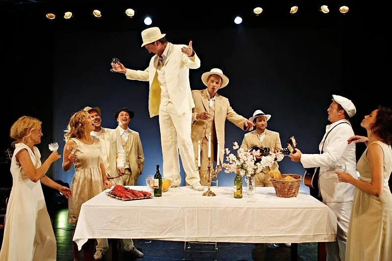 Actors performing a scene on stage dressed in vintage style clothing, with a table set with a floral centerpiece, bottles, and food, under stage lights.