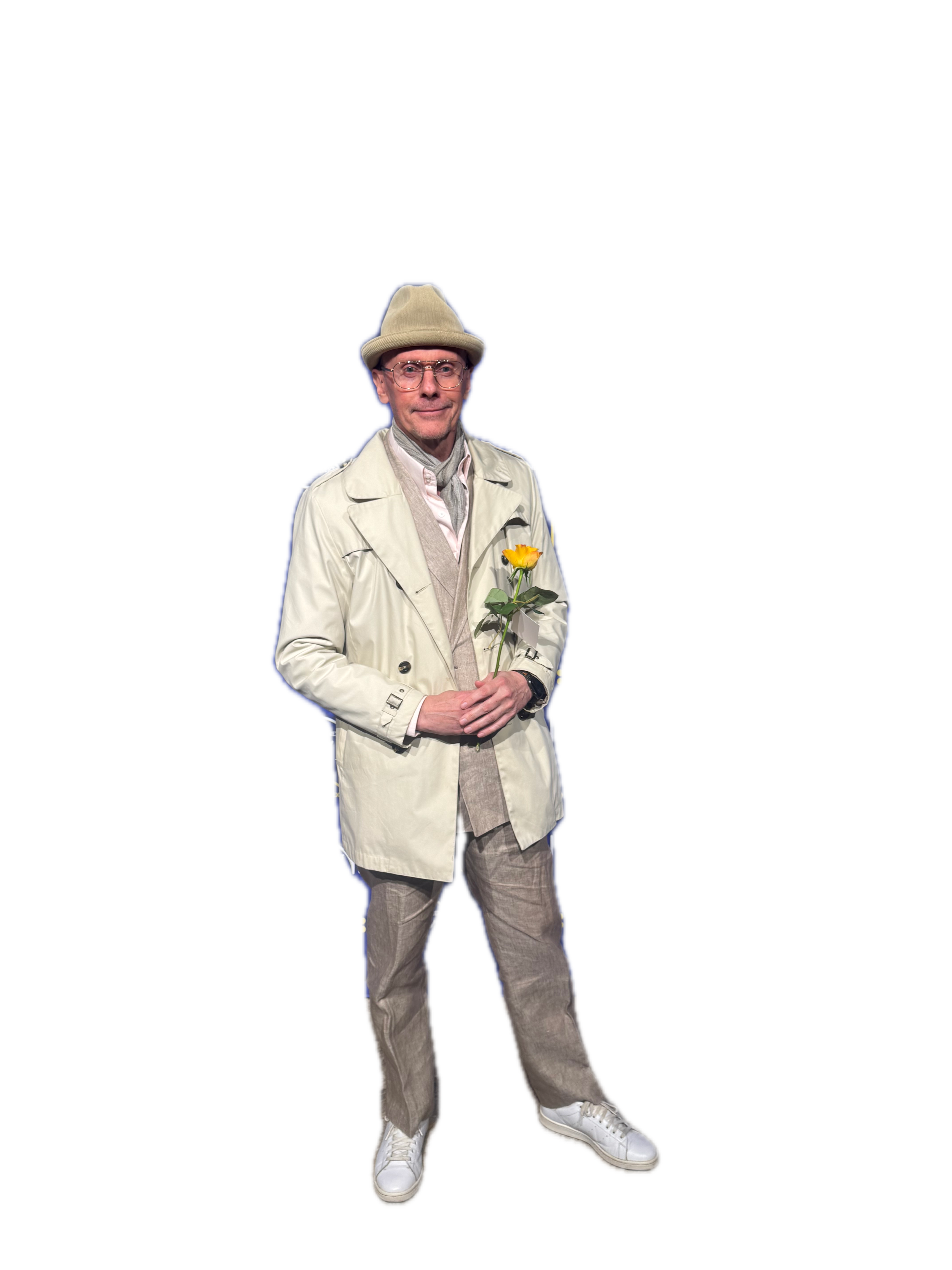 A man wearing a beige hat, glasses, a light beige trench coat, gray pants, and white sneakers is holding a yellow rose and standing against a dark background with colorful horizontal lines.